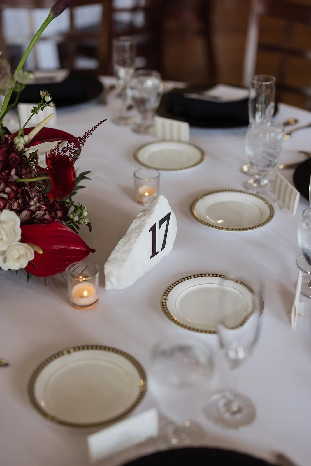 Elegant dining table decorated for a special occasion with a floral centerpiece, candles, and dinnerware. The table has place settings with white plates with gold and black borders, water glasses, and black napkins. There is a table number sign with 