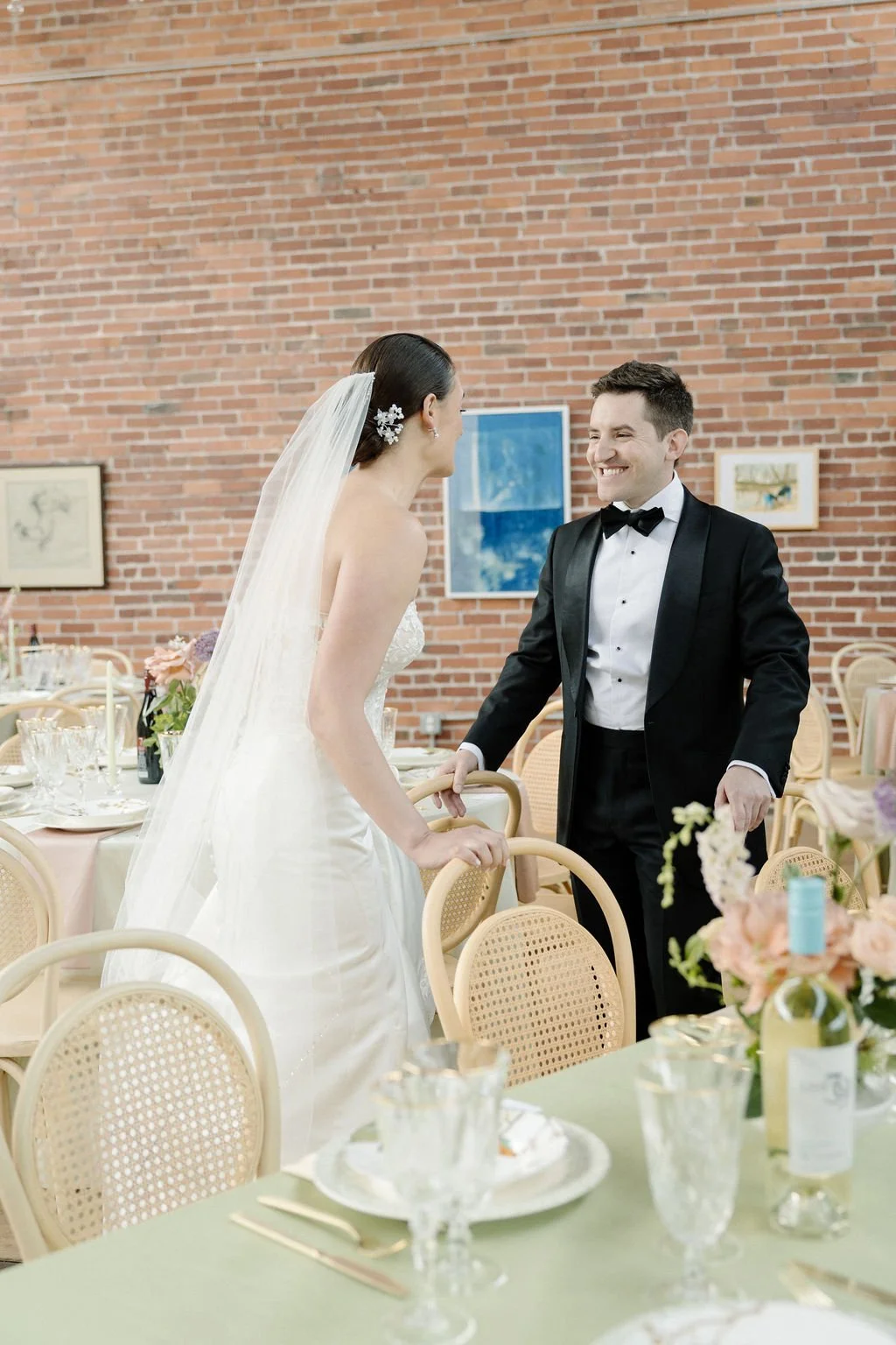 Bride and groom smiling and holding hands at wedding reception with tables and floral arrangements