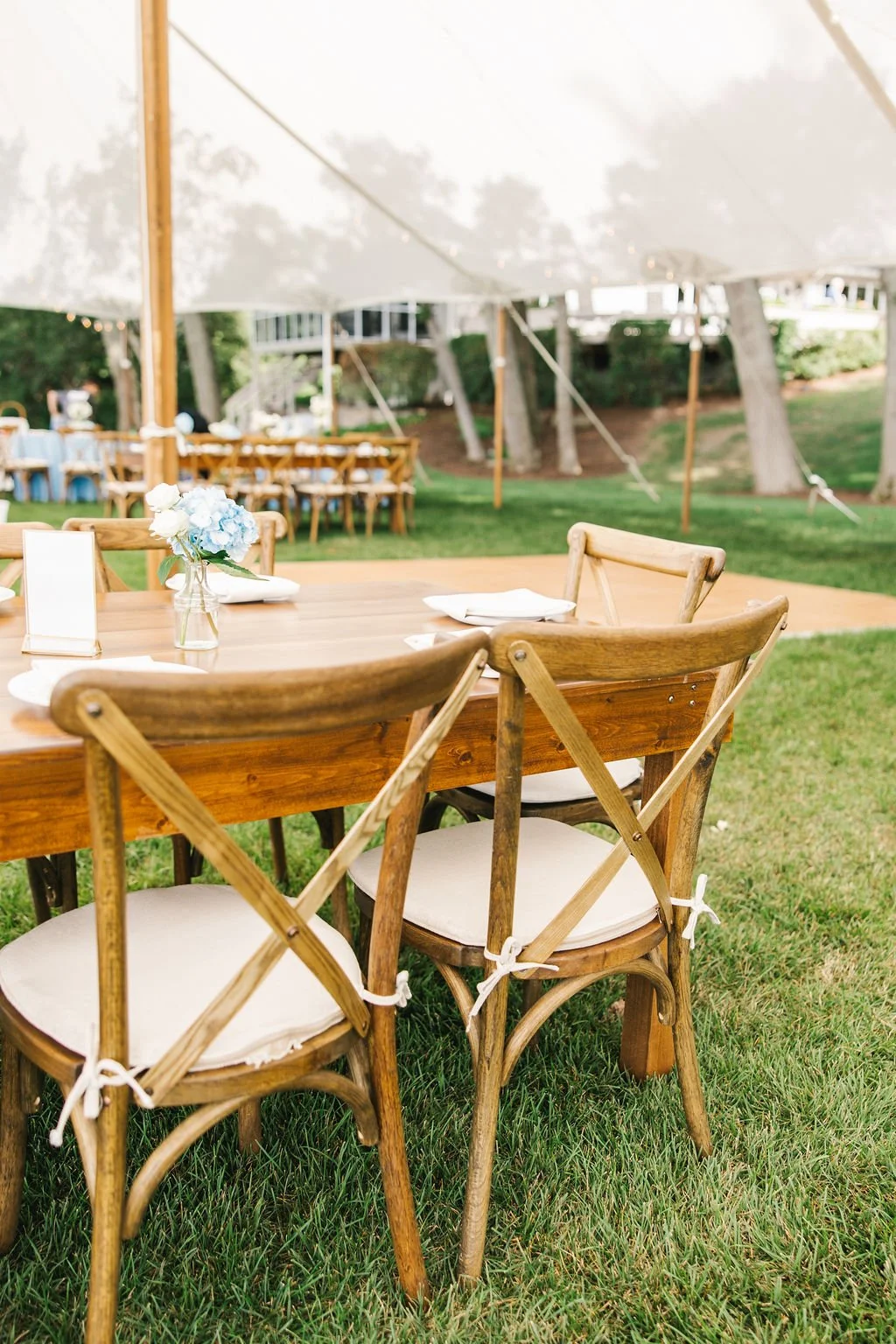 Outdoor wedding reception setup with wooden tables and chairs under a white canopy, decorated with flower centerpieces, on a grassy lawn with trees in the background.