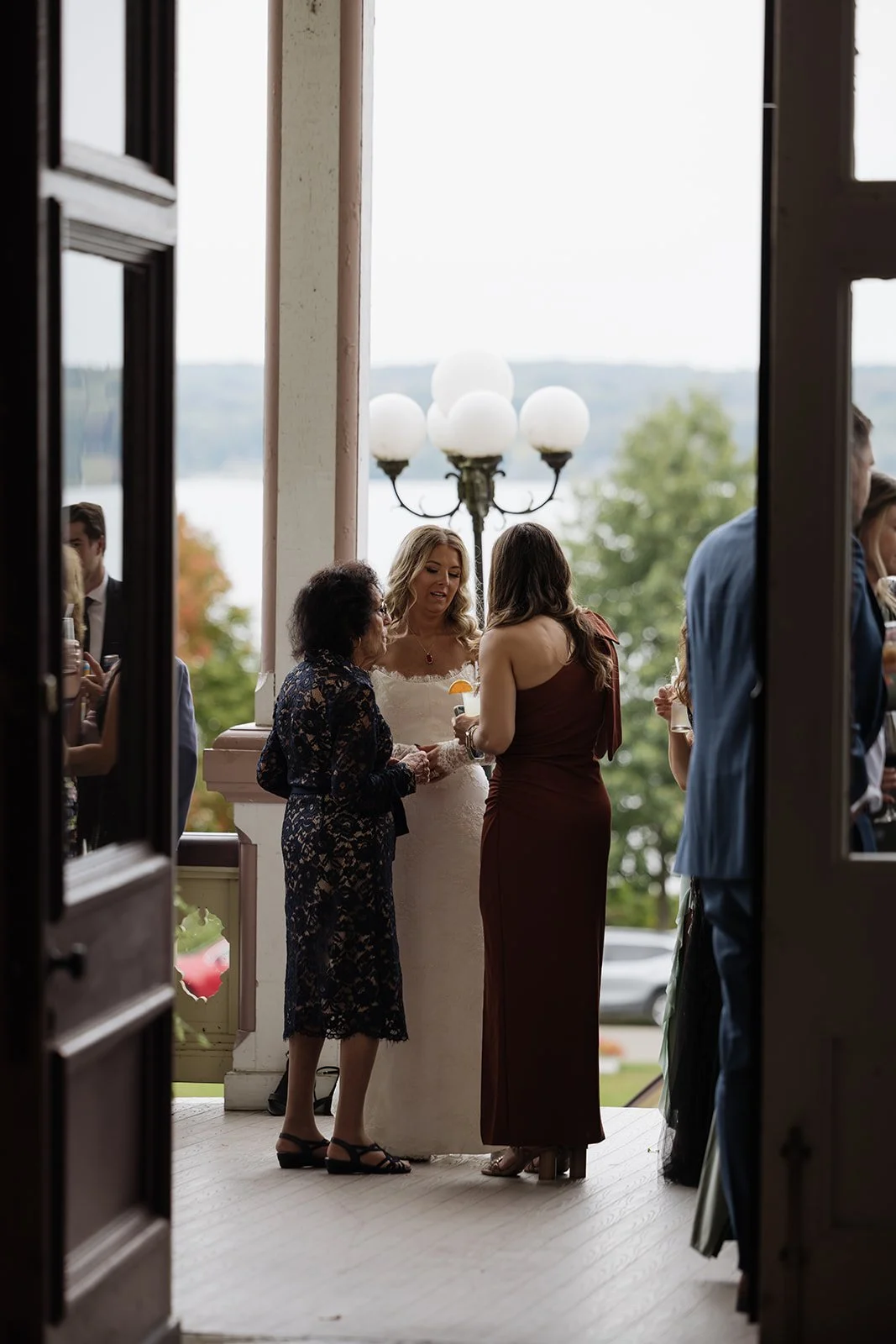 People gathered on a porch during a wedding reception, with a woman in a white dress talking to two women, one in a lace dress and the other in a sleeveless gown, holding drinks.