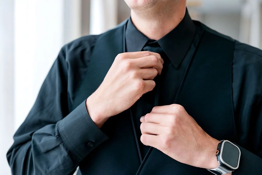 A man in a black shirt adjusts his collar or tie, wearing a smartwatch on his left wrist.