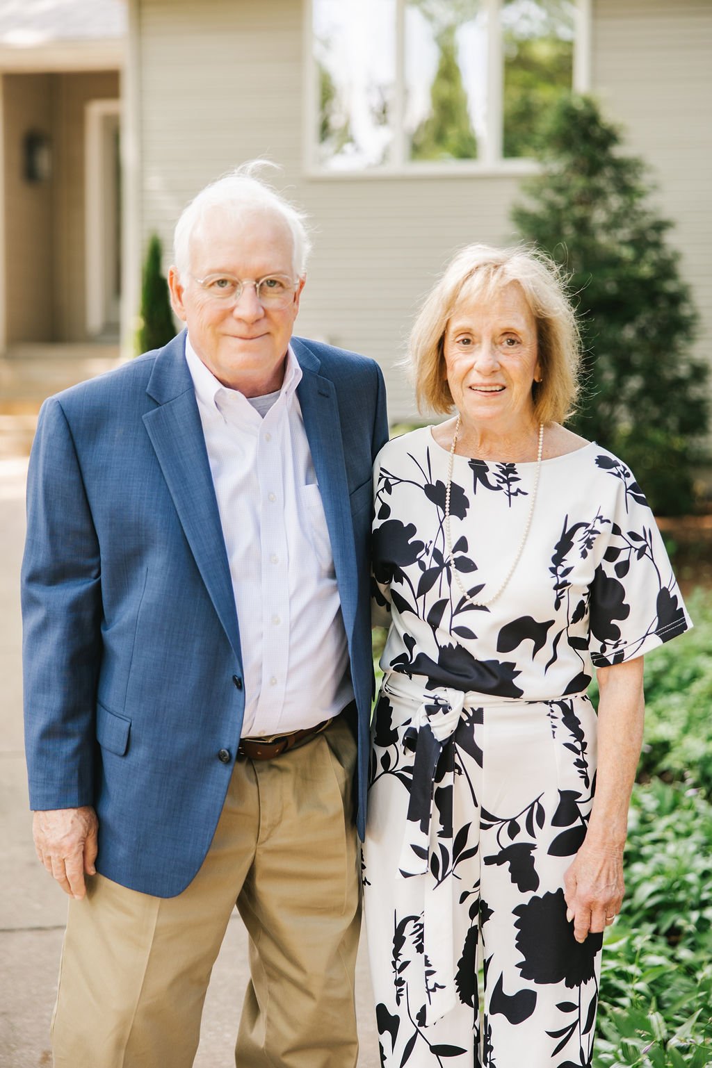 An elderly man and woman standing outside a house, smiling at the camera. The man is wearing glasses, a blue blazer, and khaki pants. The woman is wearing a white dress with black floral patterns and a pearl necklace.