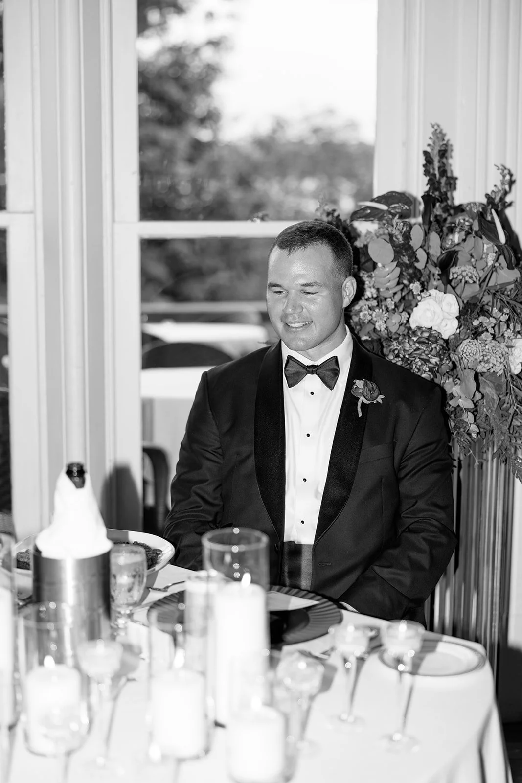 A man in a tuxedo smiling at a wedding reception, sitting at a decorated table with candles and glasses, outdoors near a window with flowers in the background.