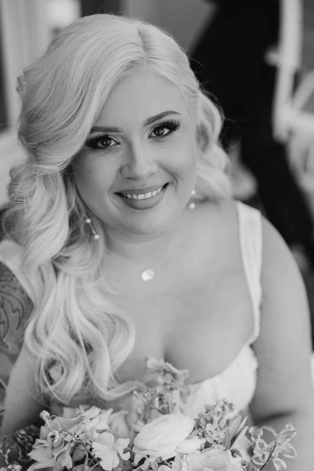 Black and white photo of a smiling woman with long wavy hair, wearing earrings, a necklace, and a dress, holding a bouquet of flowers.