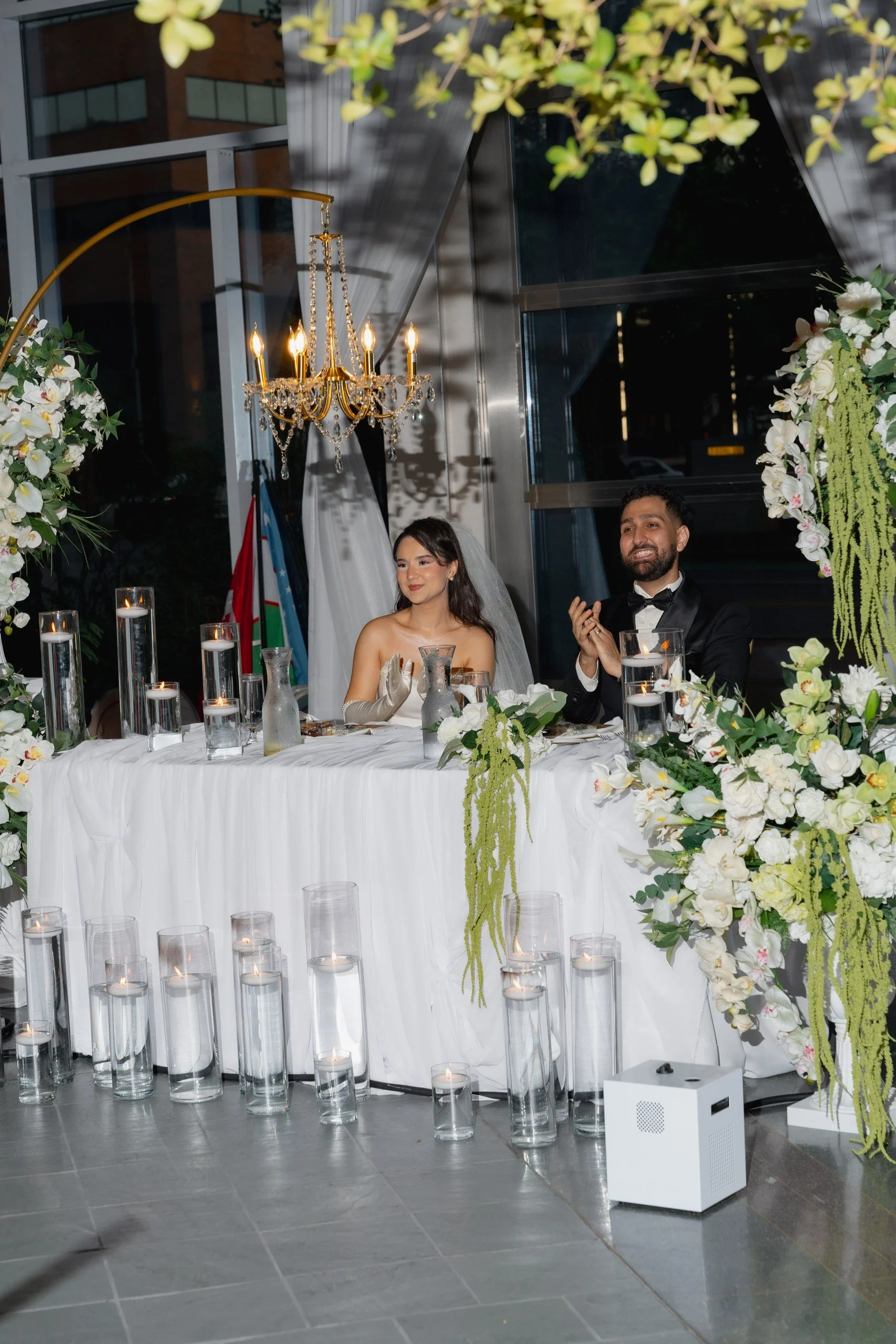 A wedding reception scene with a bride and groom sitting at a decorated table adorned with white flowers and candles. The bride is wearing a white dress and veil, while the groom is in a black tuxedo with a bow tie. There are large glass candle holde