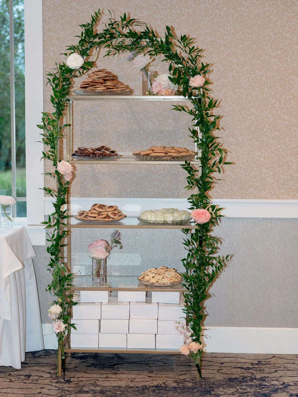 A decorated stand with flowers and cookies, featuring five shelves with cookies and treats, surrounded by pink and white flowers and greenery, located indoors near a window.