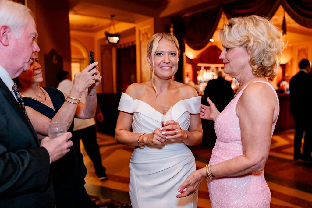 Four people at an indoor social event, two women in conversation, one woman in a white off-shoulder dress holding a drink, the other woman in a pink sequin dress, and a man taking a photo.