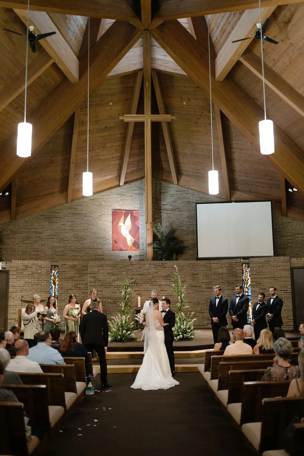A wedding ceremony taking place in a church with wooden vaulted ceiling, brick walls, and stained glass windows. The bride and groom are standing in the center, surrounded by wedding party members and guests.