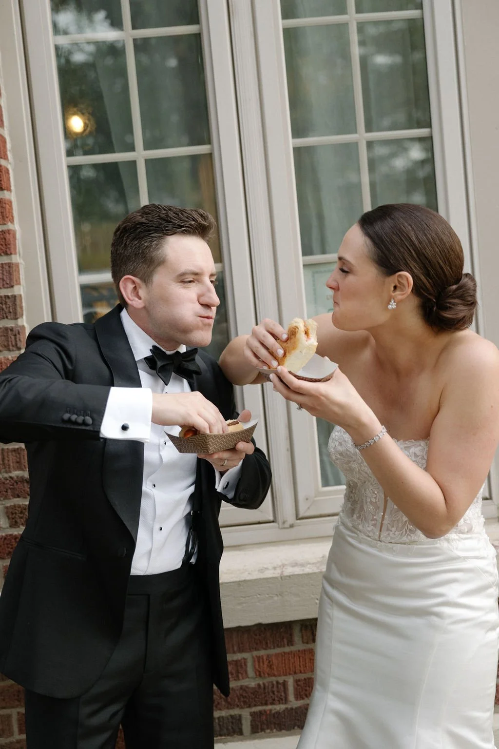 A bride and groom in wedding attire sharing a piece of cake outdoors by a window.