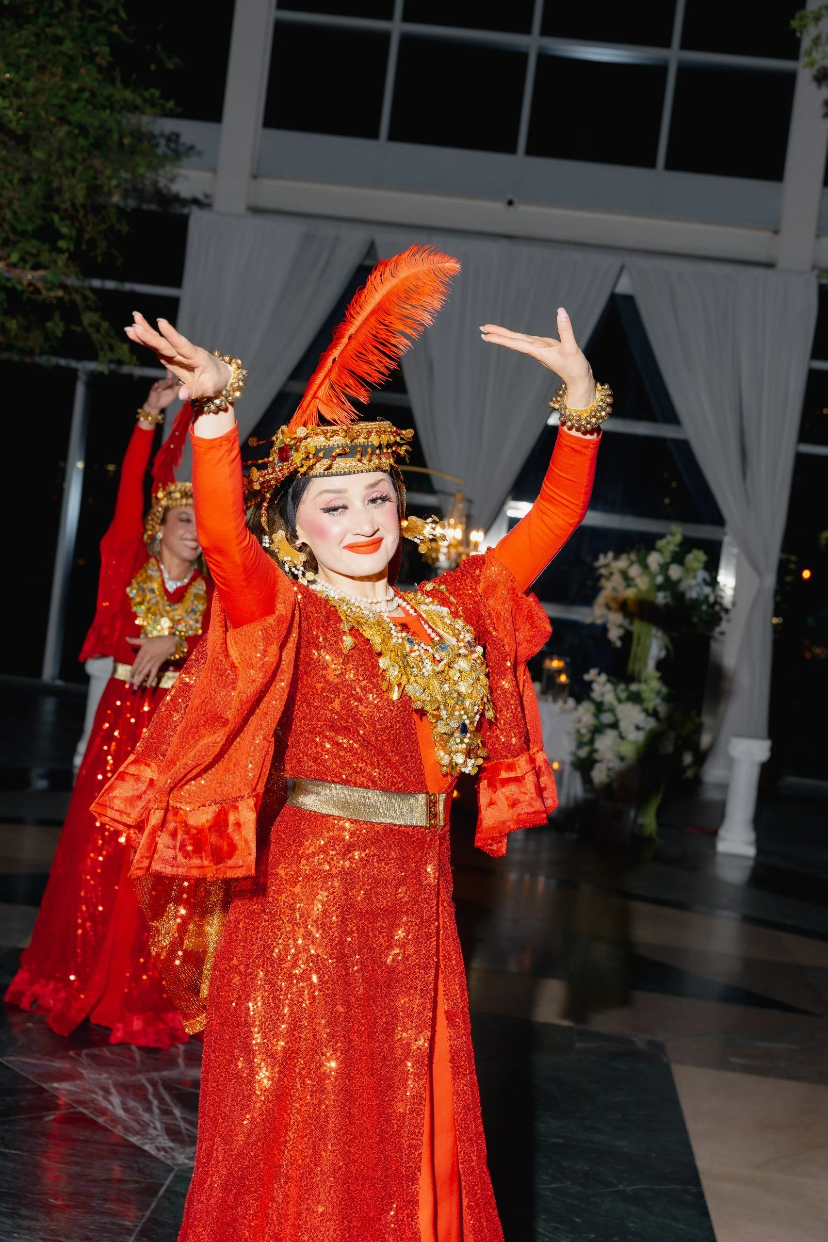 Women dancing in traditional red and gold costumes with elaborate headpieces, adorned with gold jewelry, in an indoor event setting.