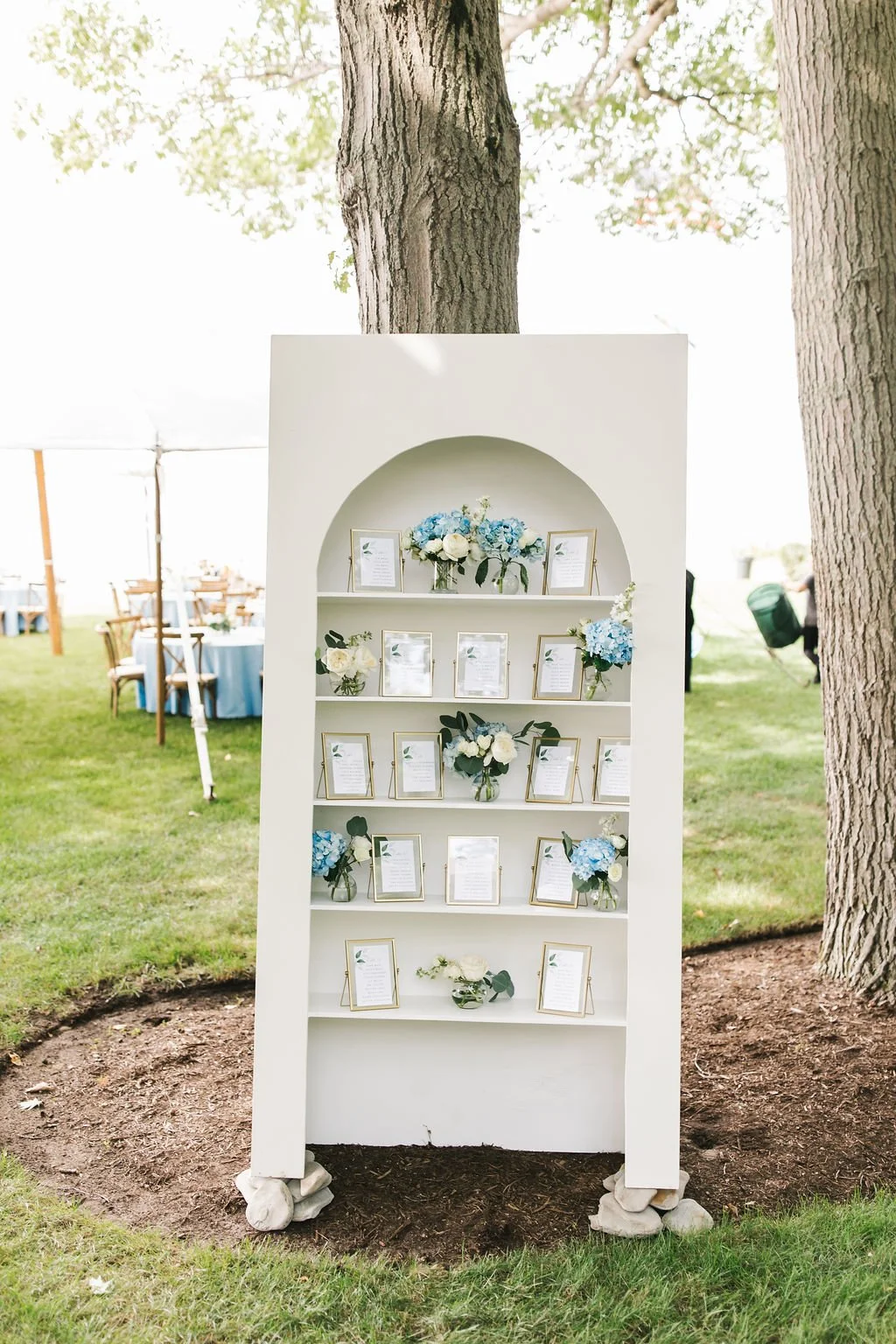 A white display shelf with framed pictures and blue and white floral arrangements, set outdoors on grass with trees in the background.