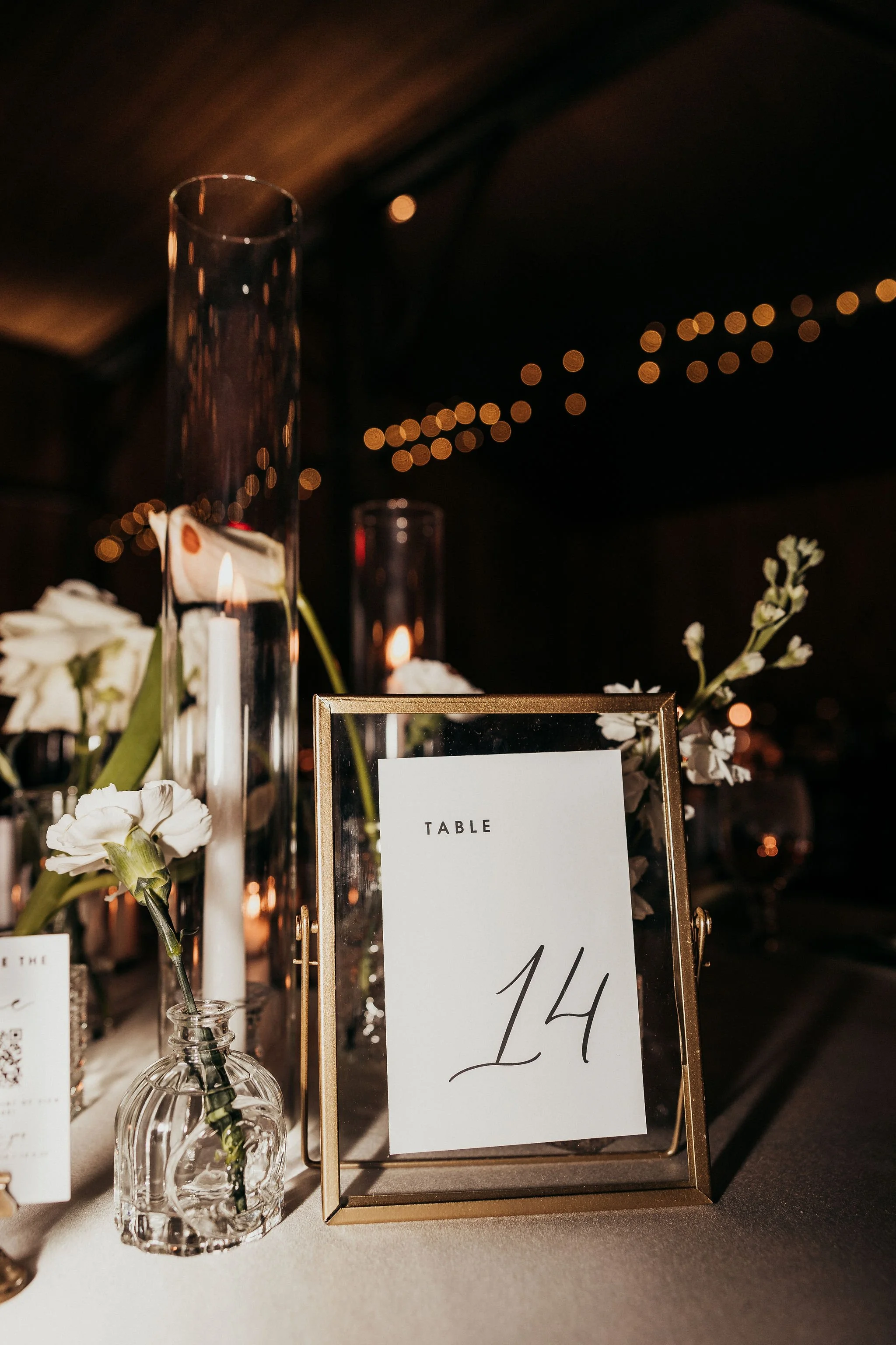 A table centerpiece with white flowers in glass vases, candles, and a framed table number sign reading '14' at an event, with string lights visible in the background.