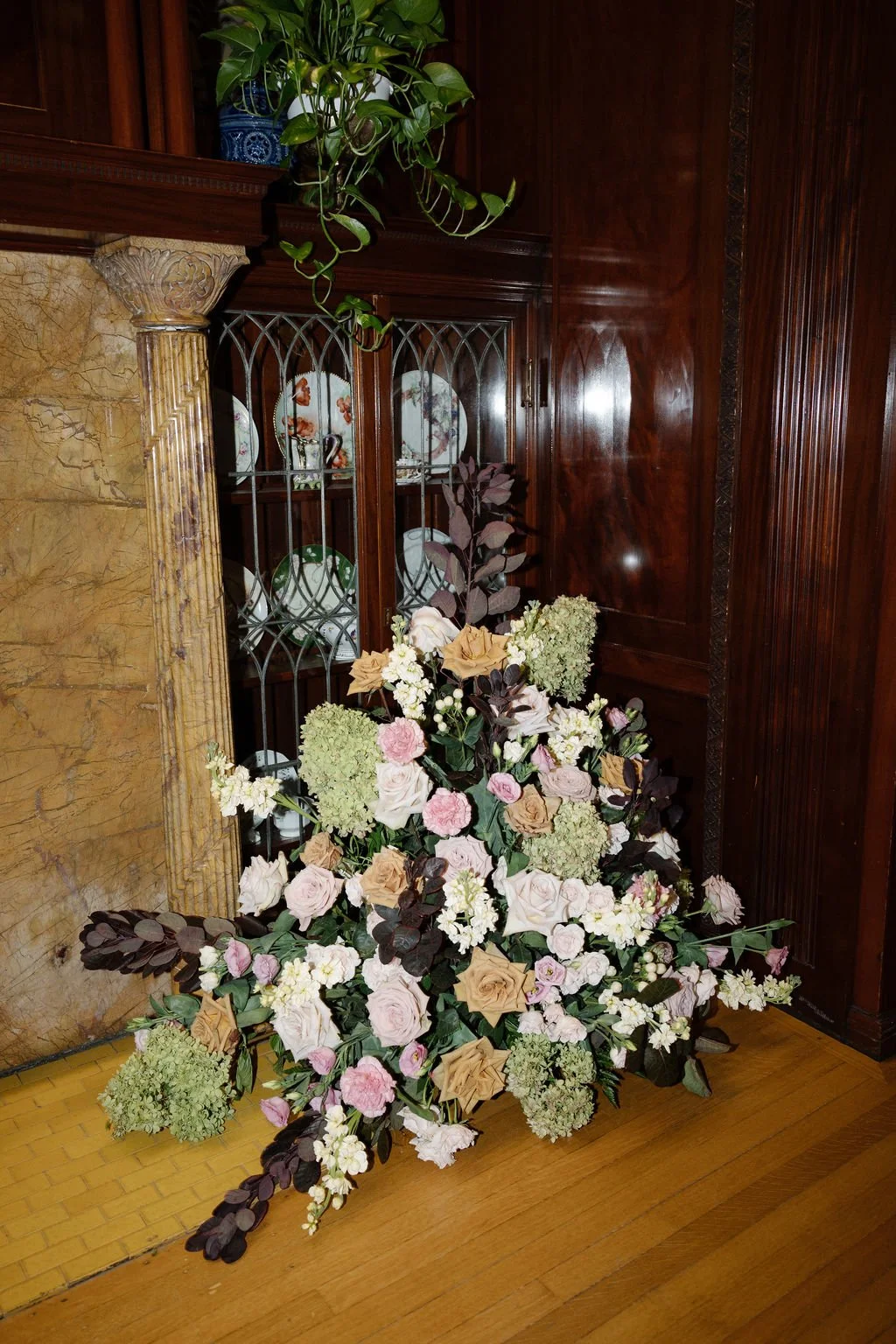 A floral arrangement with pink, white, and green flowers and dark leaves is placed on a hardwood floor next to a wooden and marble fireplace and a glass-fronted wooden cabinet displaying decorative plates in a richly decorated room.