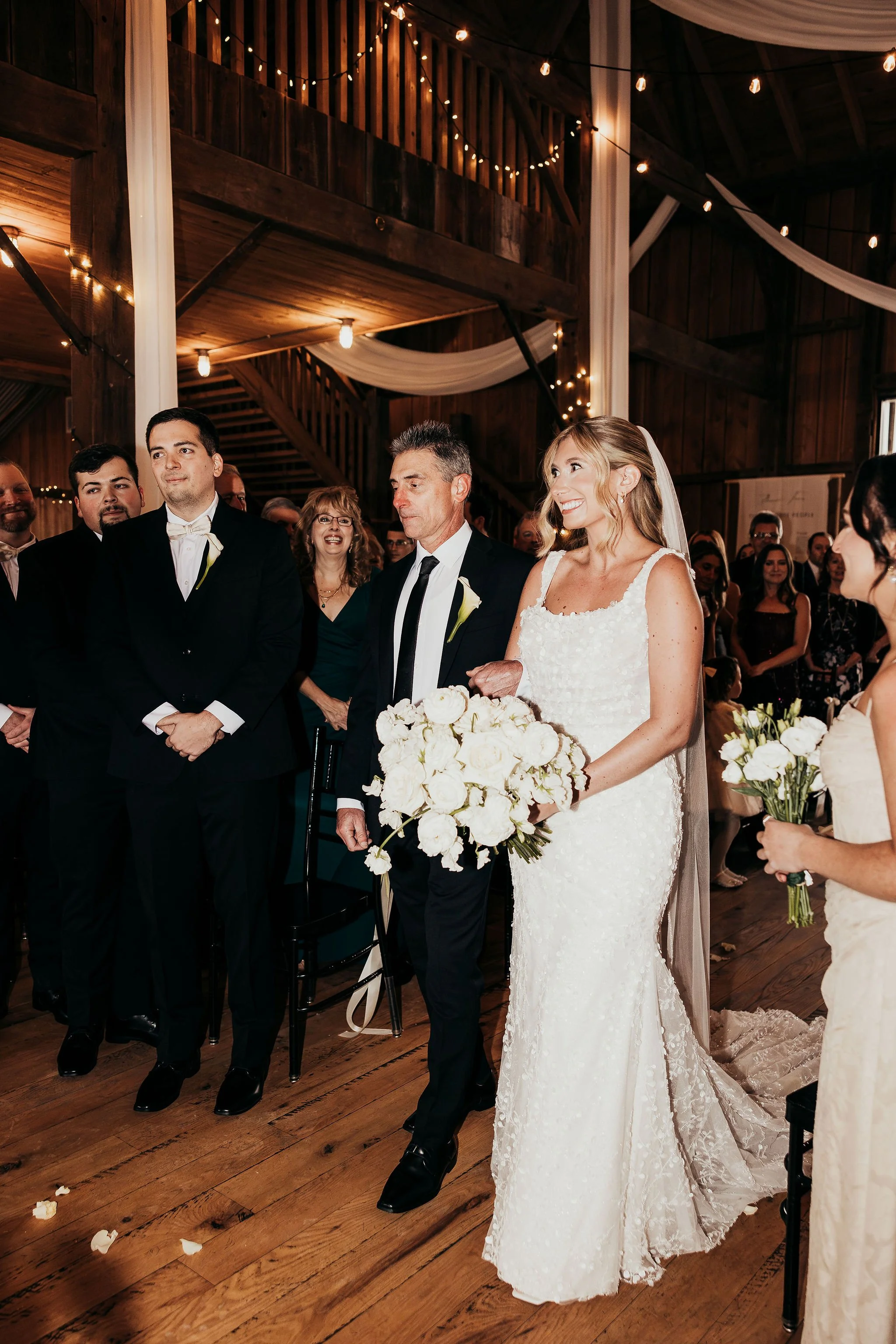 A bride in a white lace wedding gown holding a bouquet of white roses, standing next to her father who is dressed in a black suit and tie, during a wedding ceremony inside a wooden venue decorated with string lights and draped fabric.