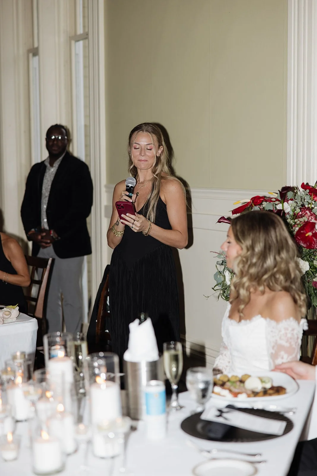 A woman in a black dress is giving a speech at a wedding reception, holding a microphone in one hand and a phone in the other. She is standing beside a bride in a white dress, seated at a decorated table with flowers, candles, drinks, and plates of f