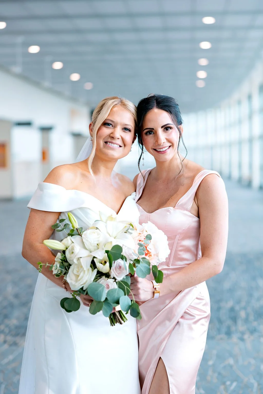 Two women, one in a white wedding dress holding a bouquet, and another in a pale pink bridesmaid dress, smiling together in a bright, modern indoor setting.