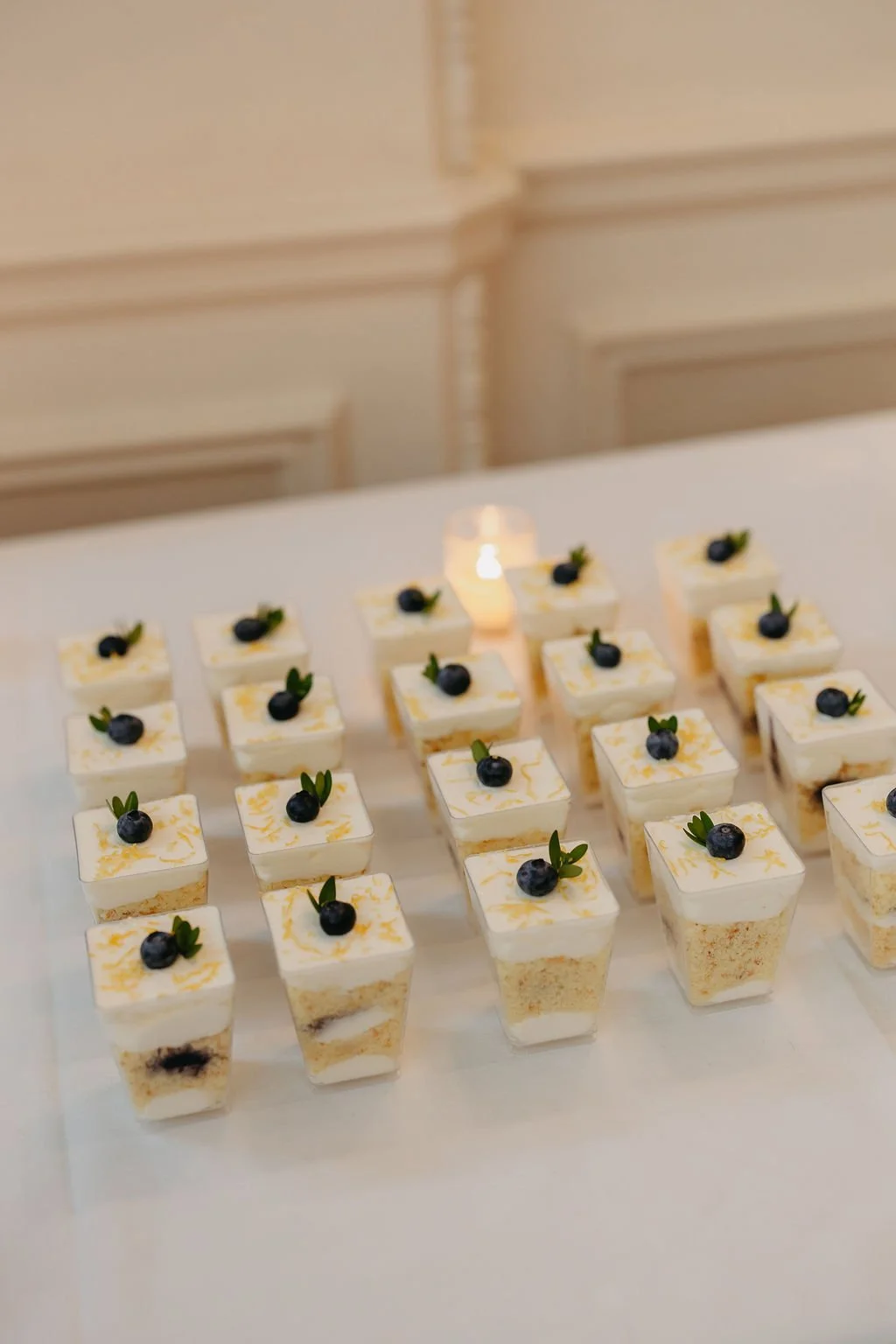 Miniature layered cakes topped with blueberries and green leaves, arranged on a white surface with a candle in the background.
