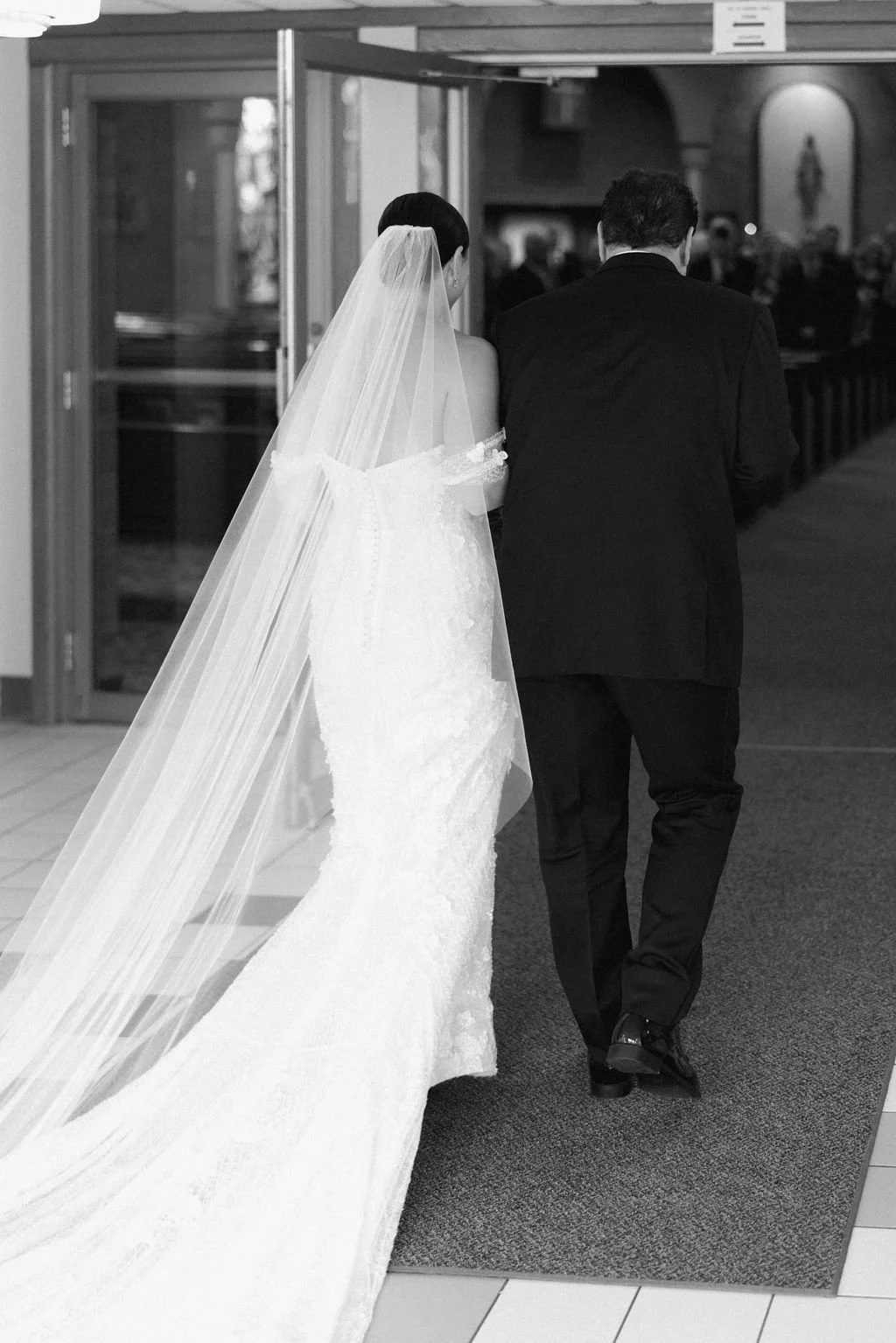 A bride in a wedding dress and veil walking with a man in a suit, likely at a wedding ceremony or reception.