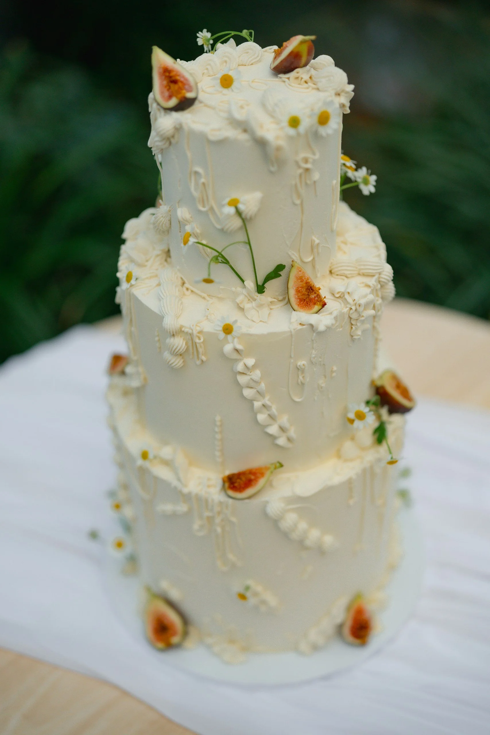 A two-tiered white wedding cake decorated with figs cut in half, small white flowers, and cream-colored piping decorations.
