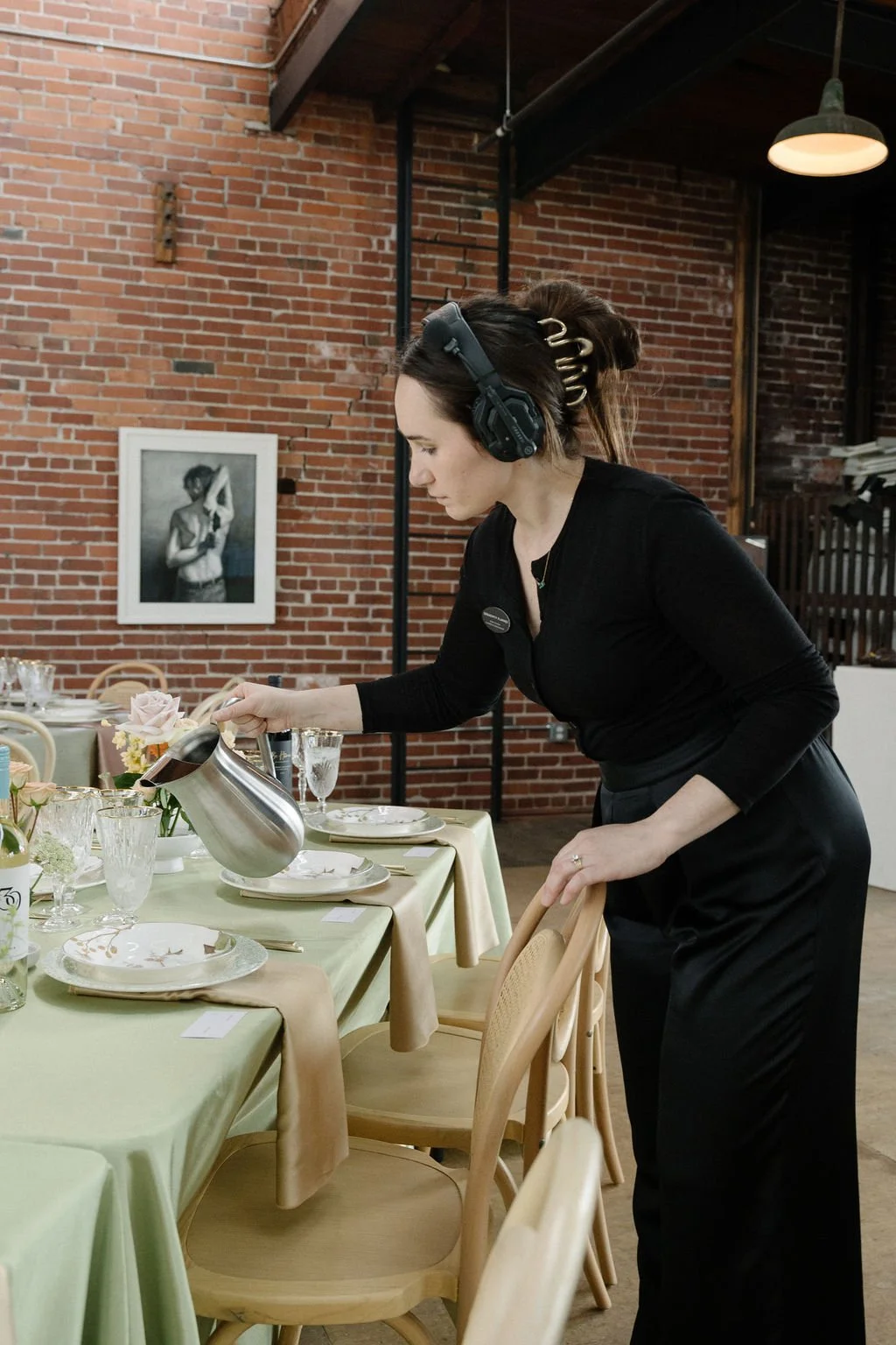 A woman with dark hair, wearing black clothing and headphones, is pouring water into a glass on a dining table set with plates, glasses, and floral arrangements in a restaurant with brick walls.