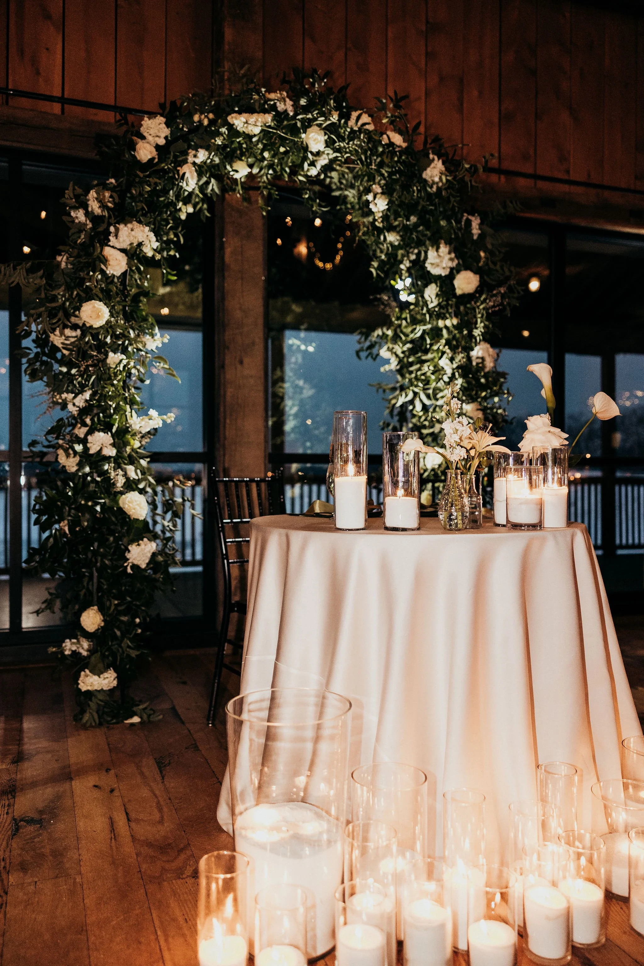 A decorated event space with a round table covered with a beige tablecloth, adorned with several candles in glass holders and a small floral arrangement. Behind the table, there is a floral arch made of greenery and white flowers, set against large w
