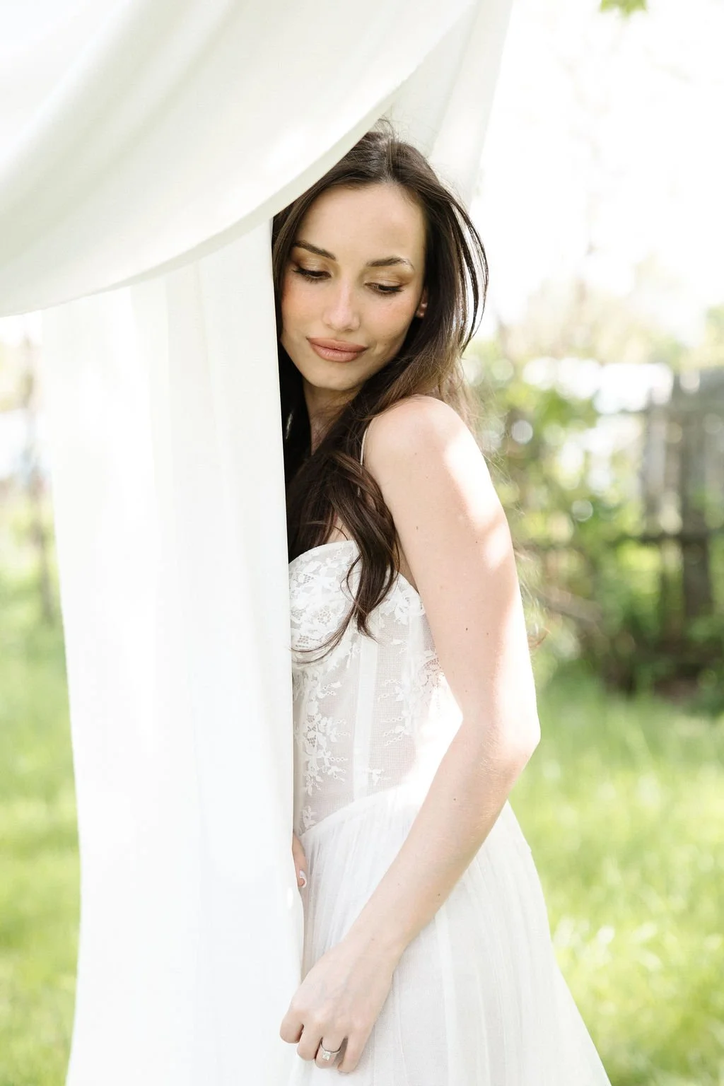 A woman with long brown hair wearing a white lace dress stands outdoors near a white curtain, with a green background of trees and grass.