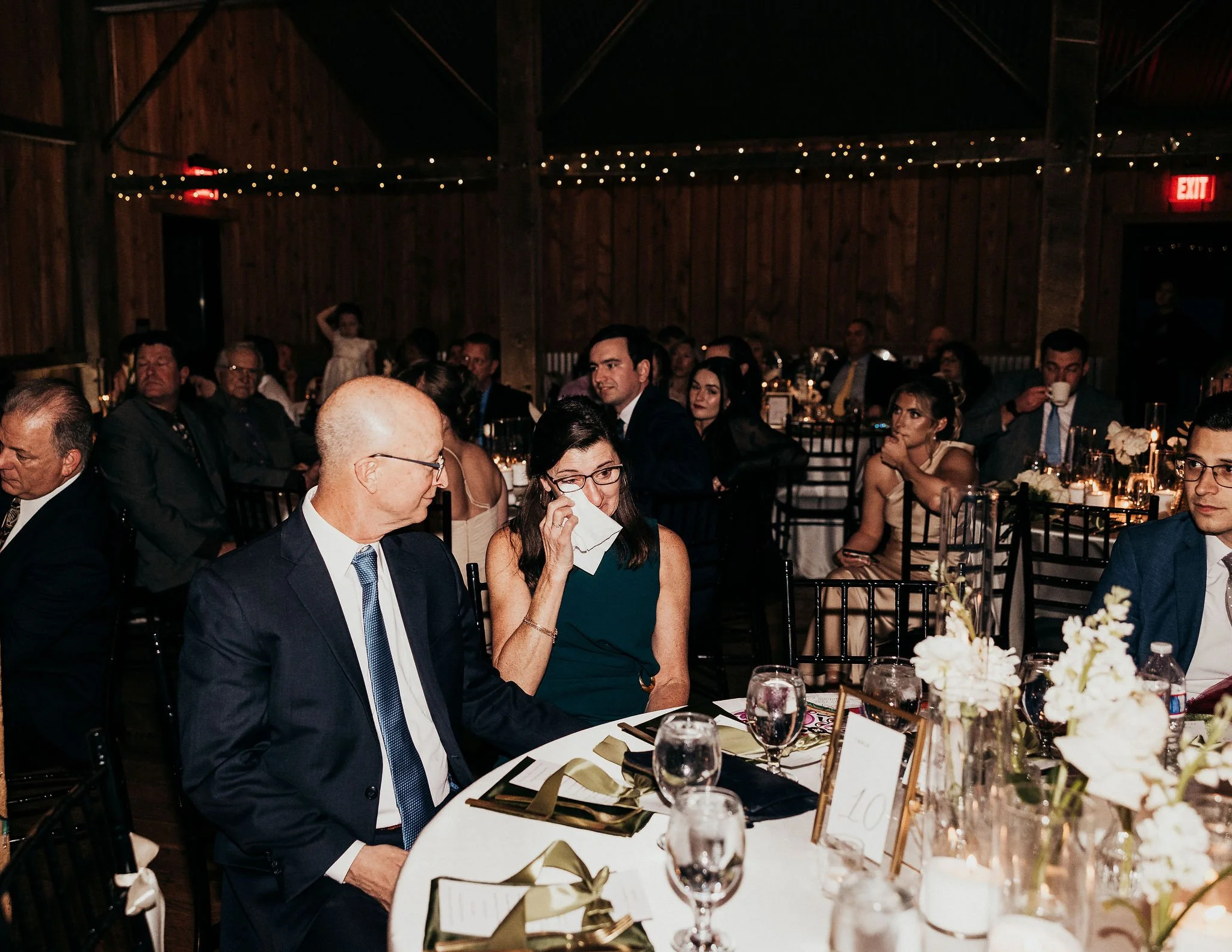 People sitting at round tables in a rustic wooden venue with string lights hanging from the ceiling during a formal event or dinner.