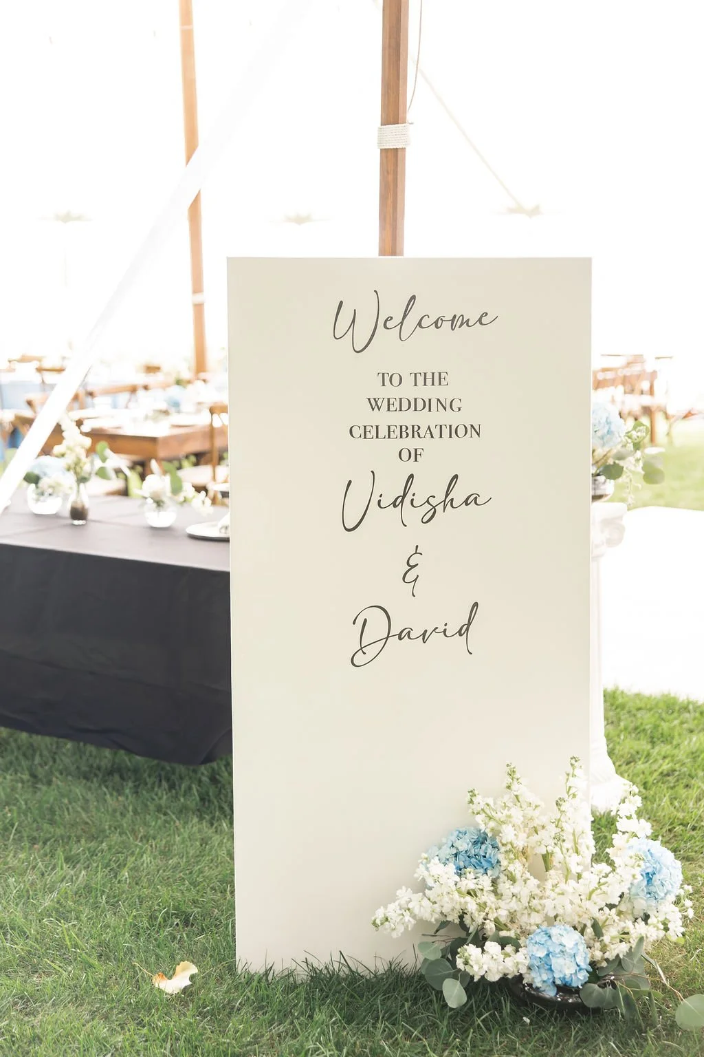 Wedding welcome sign with floral decoration at outdoor wedding reception, featuring the names Vidisha and David.