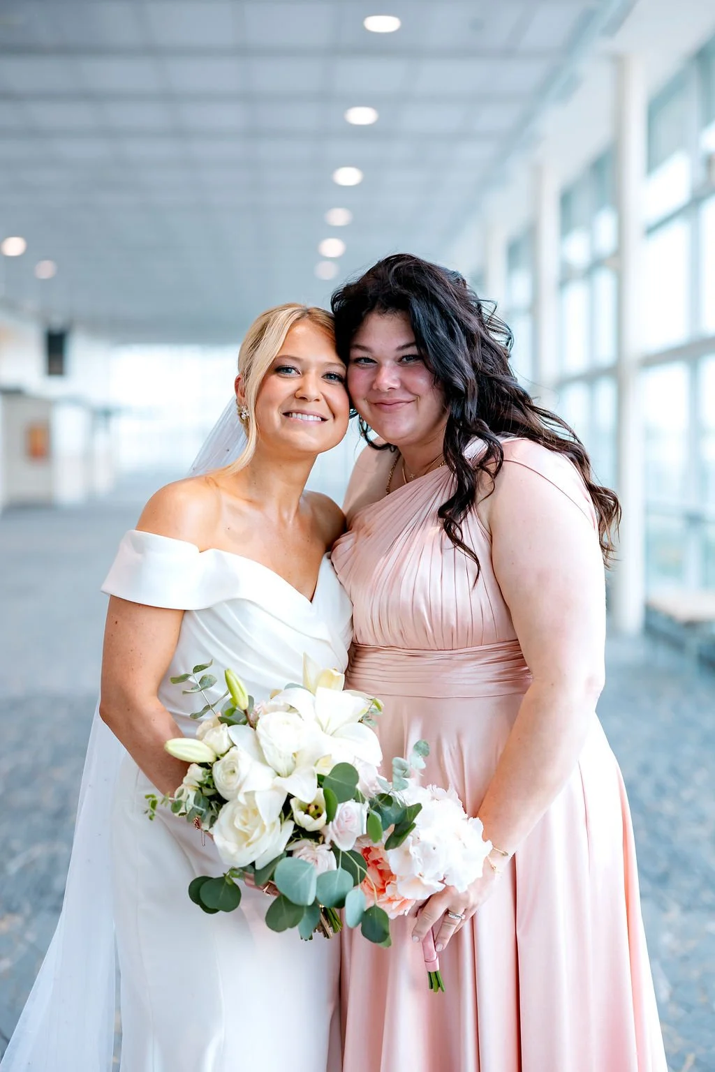 Two women, one in a white wedding dress holding a bouquet of white flowers, and the other in a pink bridesmaid dress, stand close together in a bright, modern indoor space with large windows.