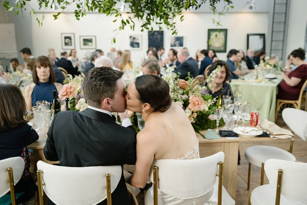 A wedding reception with the bride and groom sharing a kiss at their table, surrounded by guests and floral arrangements.