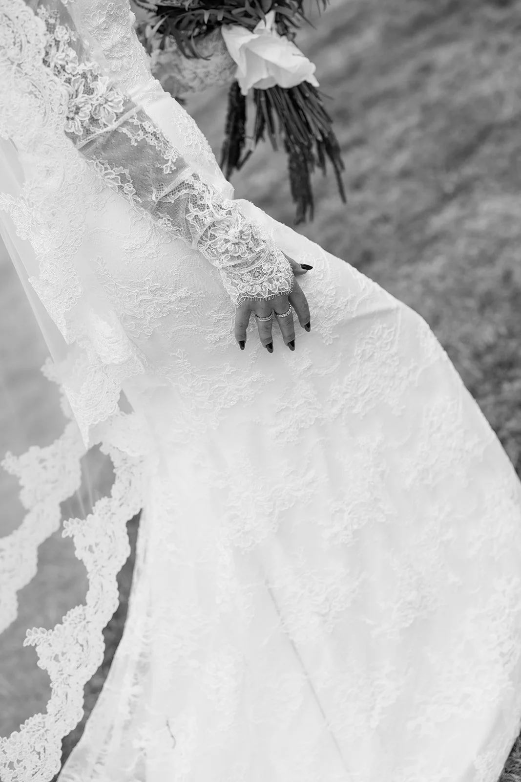 Close-up black and white photo of a bride’s lace dress, showing her hand with rings, and holding a bouquet of flowers.