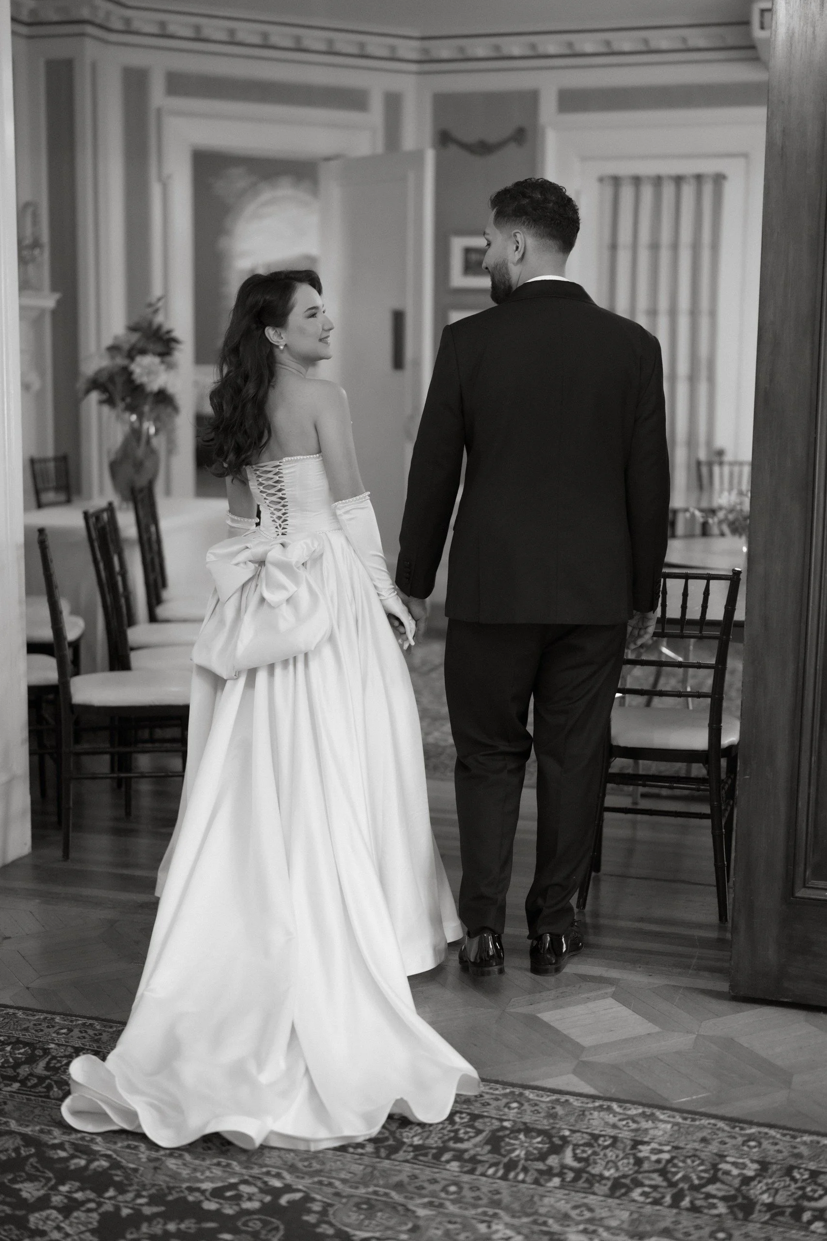 A bride and groom holding hands inside a wedding venue with chairs and floral decorations.