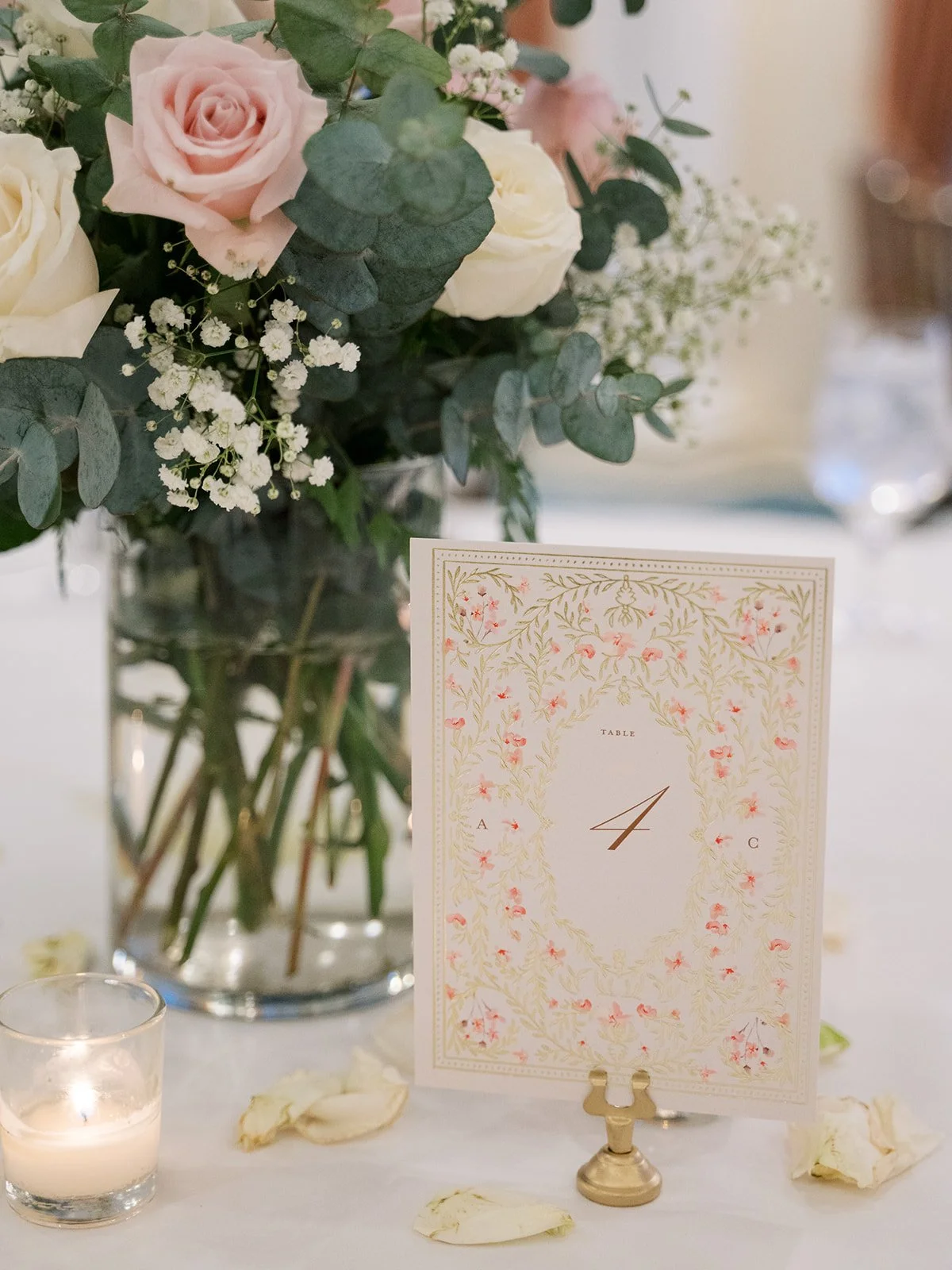 A floral centerpiece with pink and white roses, baby's breath, and eucalyptus in a clear glass vase on a table. A table number card with floral design and the number 4 is displayed on a brass holder, with white rose petals and candles surrounding it.