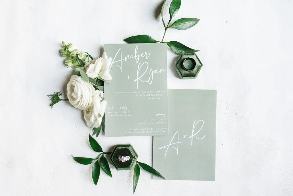 Wedding invitation suite with a light green background, white calligraphy, surrounded by white flowers, green leaves, and two green hexagonal ring boxes with engagement rings inside.
