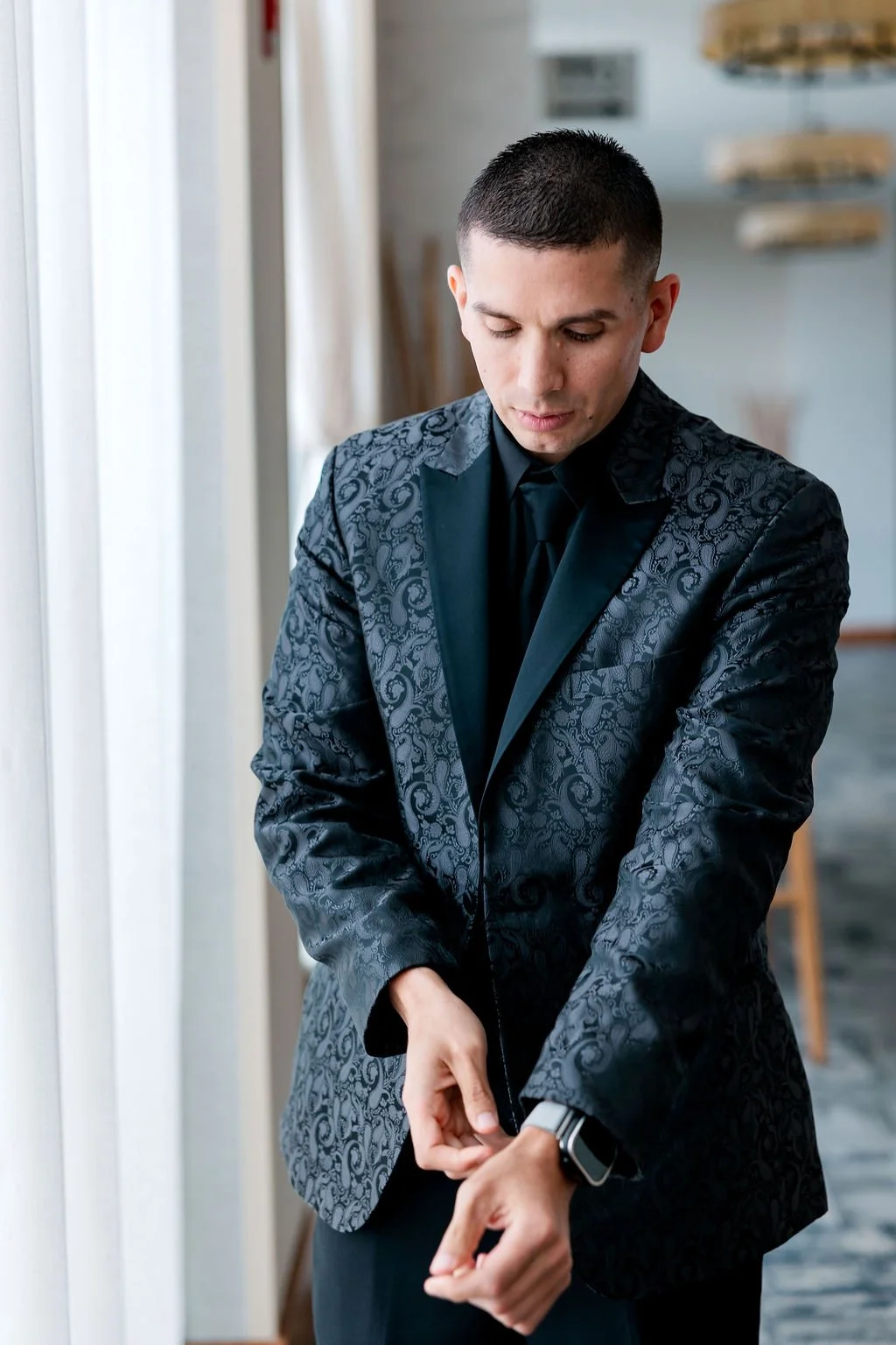 A man in a dark, patterned tuxedo is adjusting his cuff in a well-lit room.