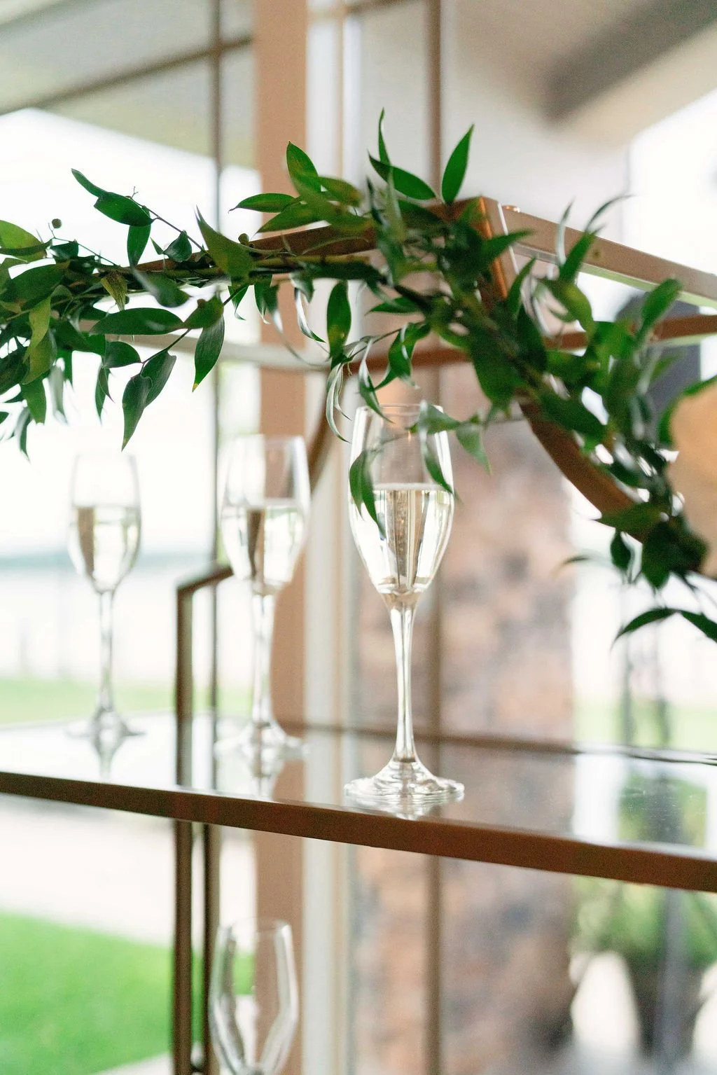 Champagne flutes and a glass vase with greenery on a reflecting surface.
