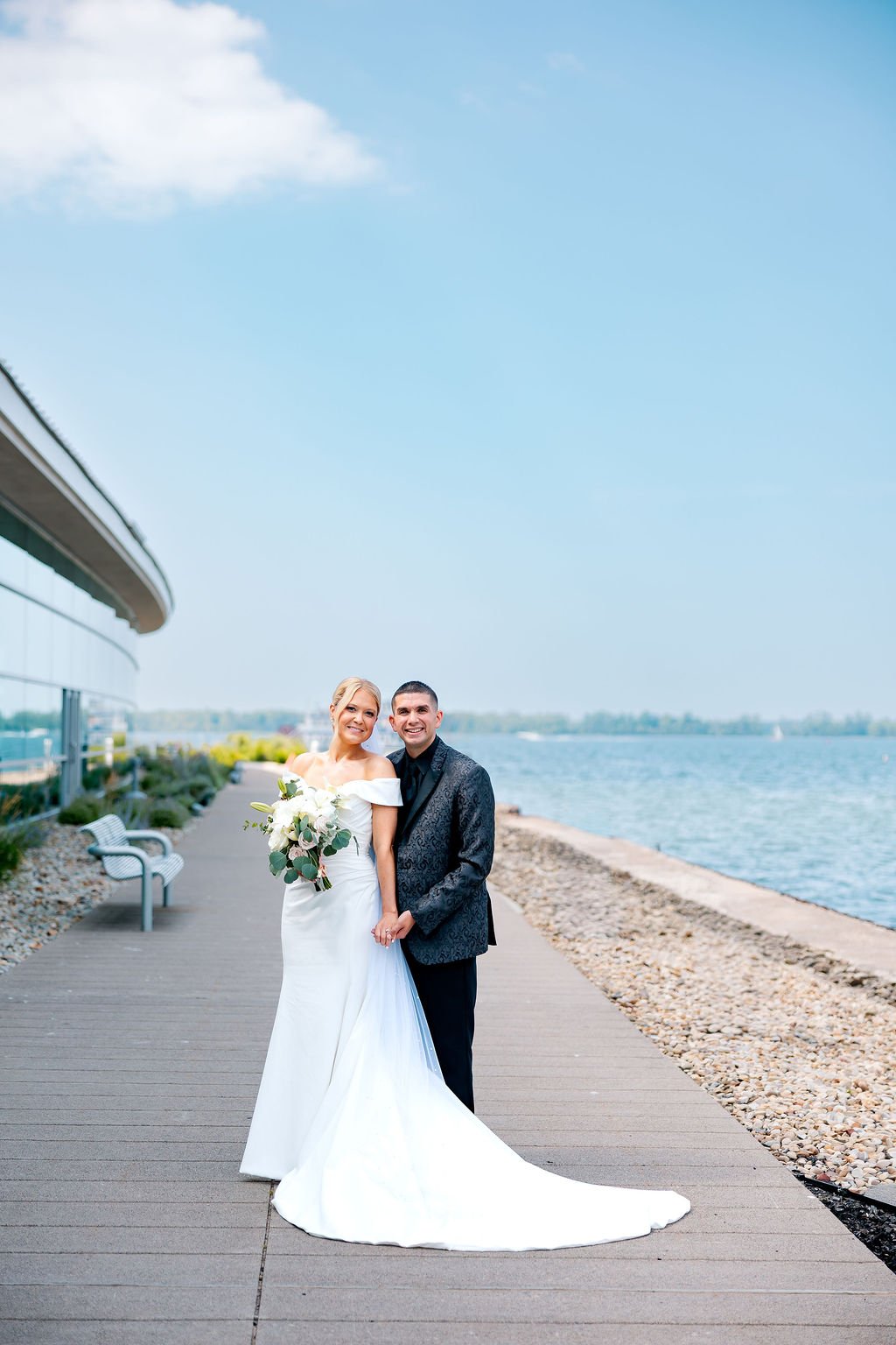 A newlywed couple stands on a waterfront promenade, smiling and holding hands, with a modern building on the left and water on the right, under a partly cloudy sky.