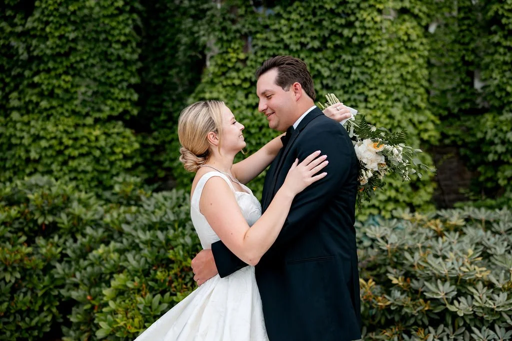 A bride and groom embracing outdoors, surrounded by greenery, with the bride holding a bouquet of flowers.