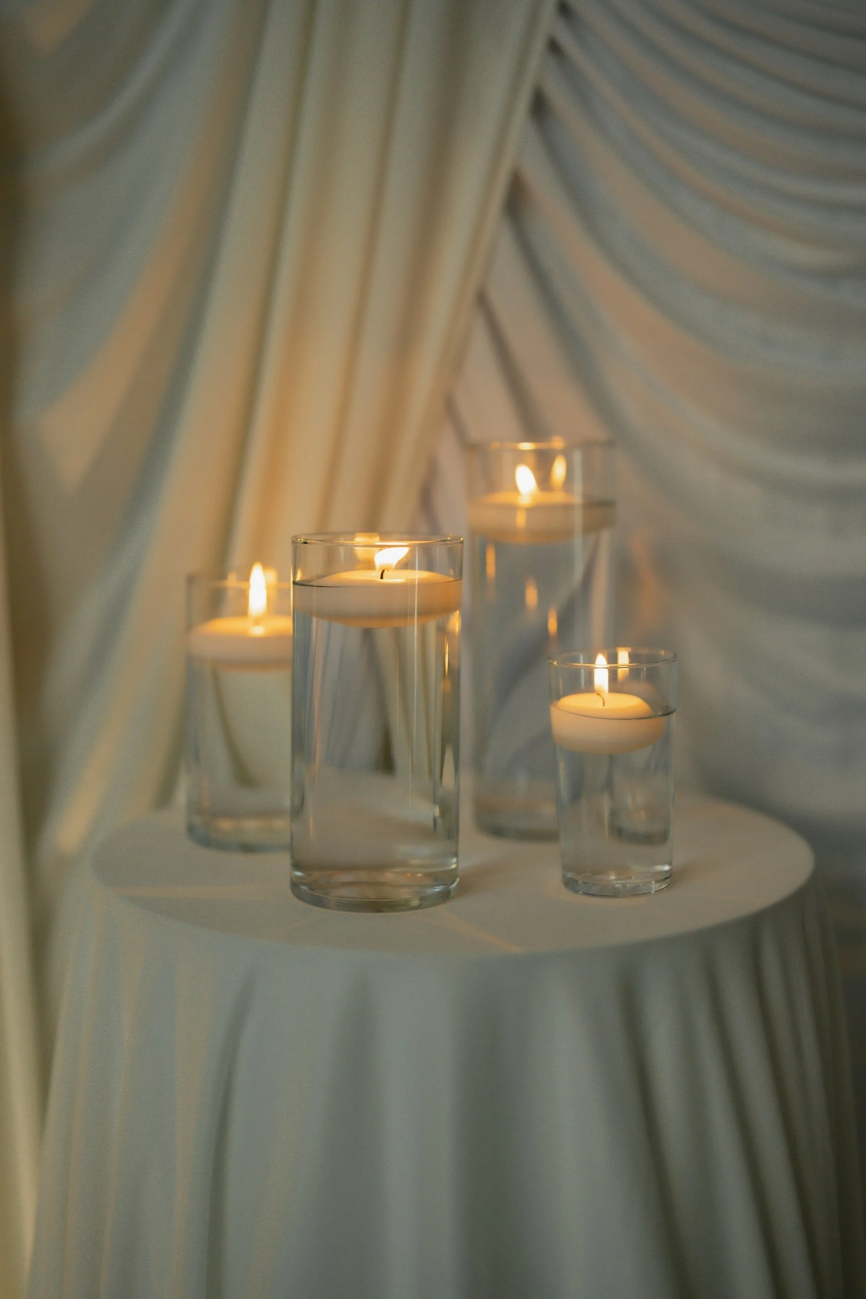 Several lit candles in glass holders on a white surface, with draped fabric in the background.