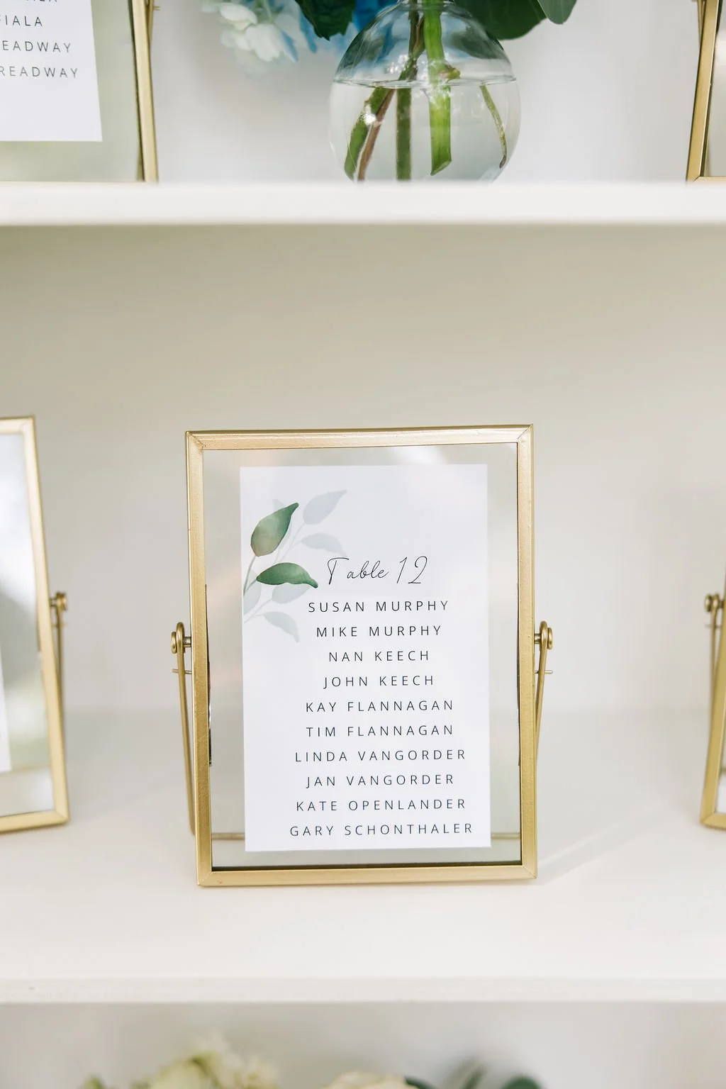 Table 12 seating chart for a wedding with names listed in black text, decorated with a green leaf illustration, framed in a gold frame.