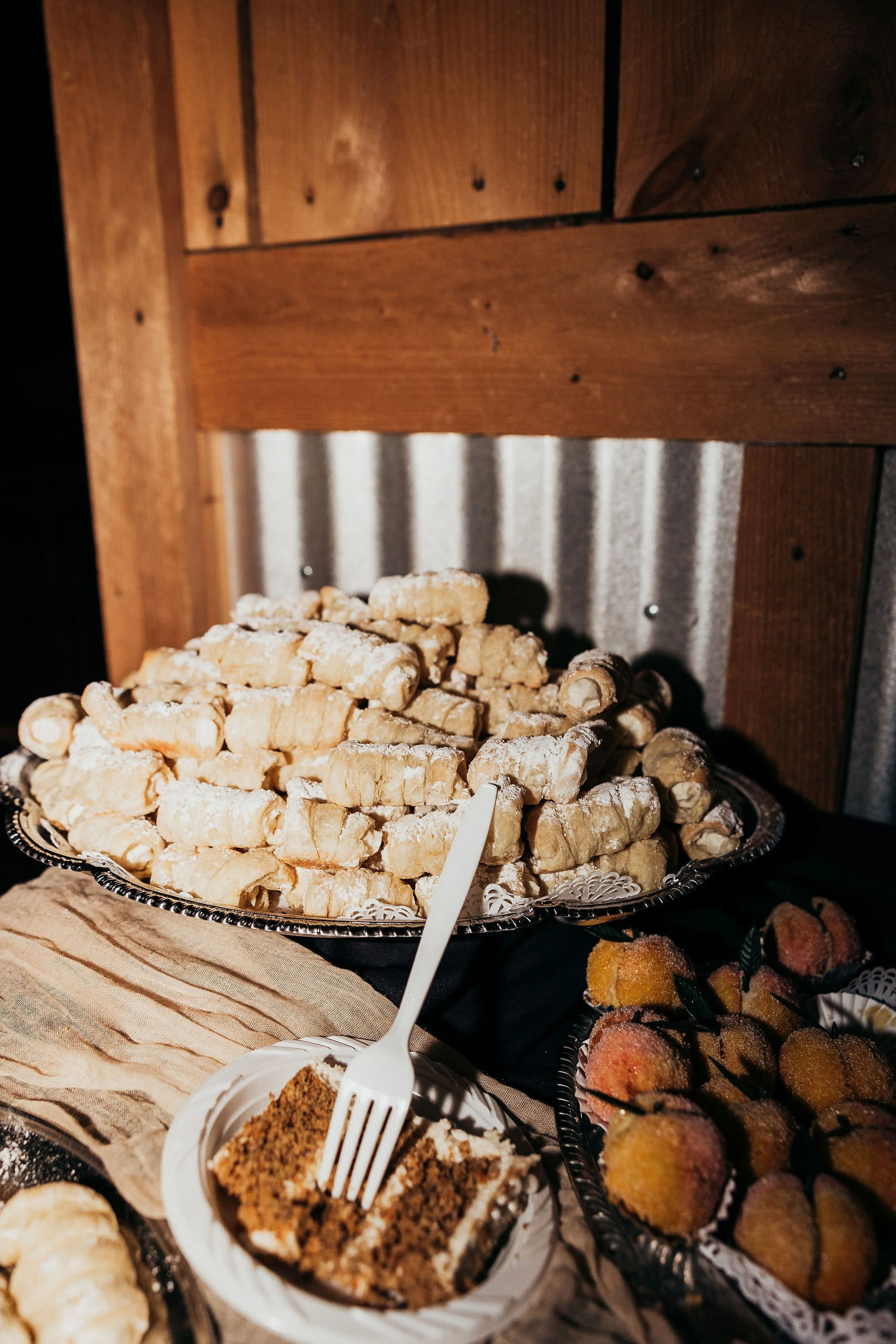 Assorted desserts on a table, including a pile of powdered cookies, a slice of layered cake, and mini cupcakes, with a wooden and metal backdrop.