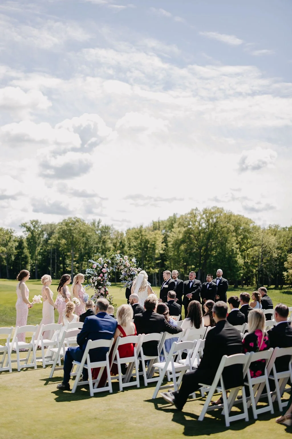 Outdoor wedding ceremony with a bride and groom standing under a floral arch, surrounded by bridesmaids in pink dresses and groomsmen in dark suits, seated guests on white chairs, lush green field and trees, cloudy sky.