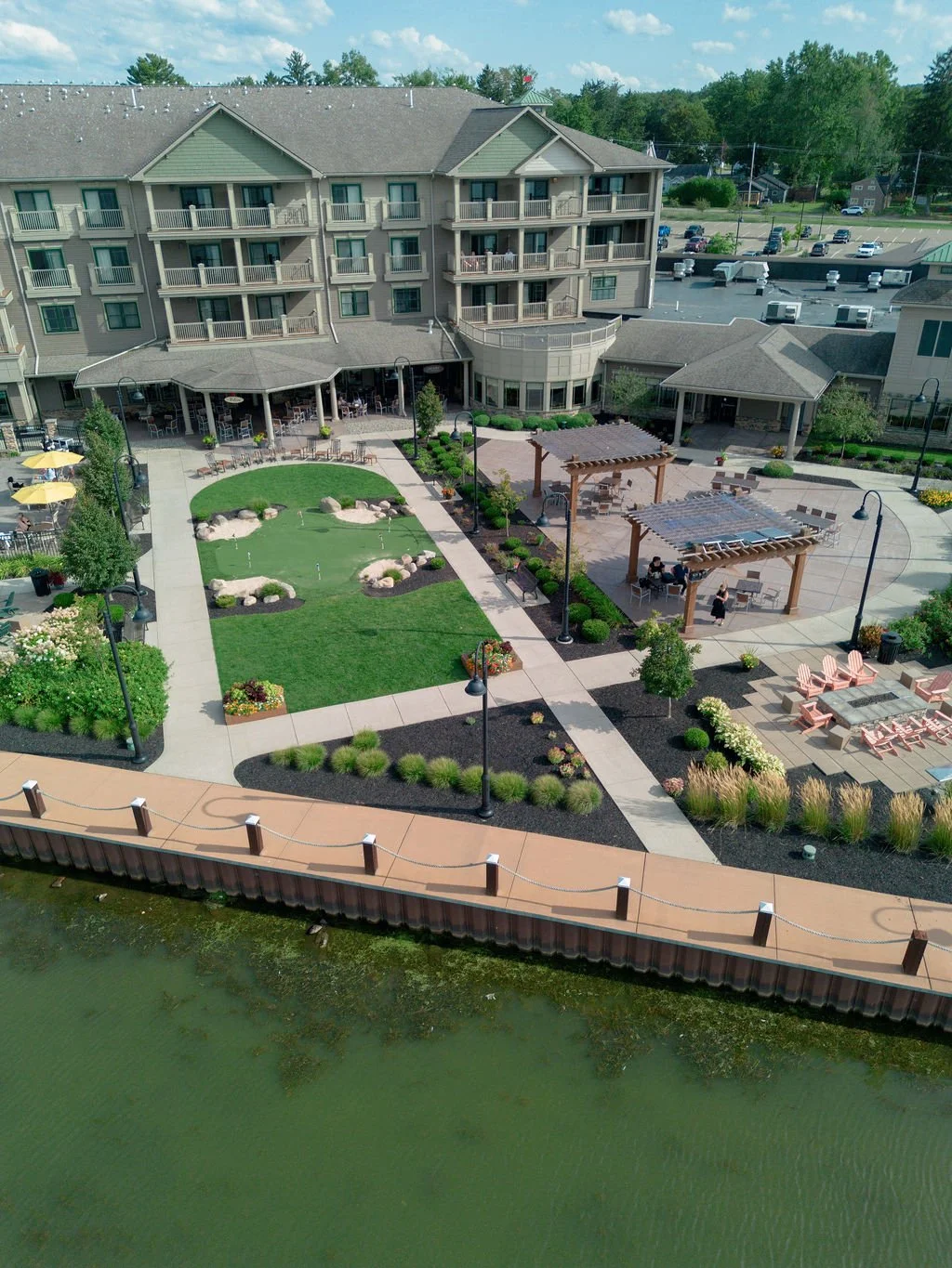 Aerial view of a landscaped courtyard with grassy areas, walking paths, outdoor seating, pergolas, and a pond with a walkway along the water, surrounded by a multi-story building and parking lot.