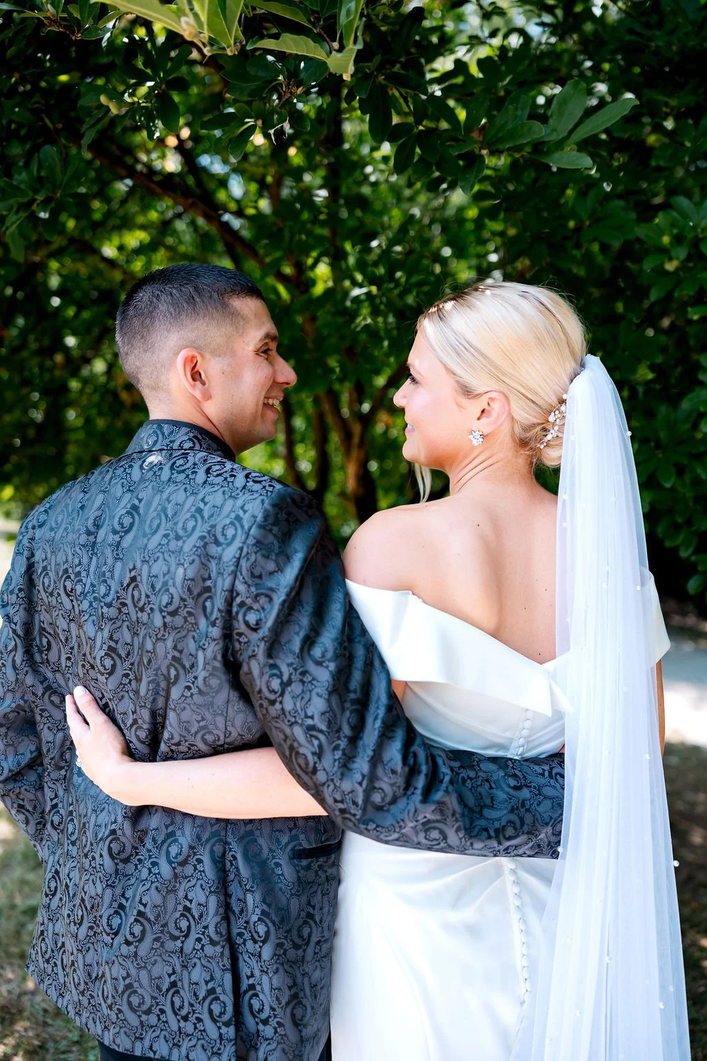 A newlywed couple in wedding attire, standing outdoors among green trees, gazing at each other and smiling.