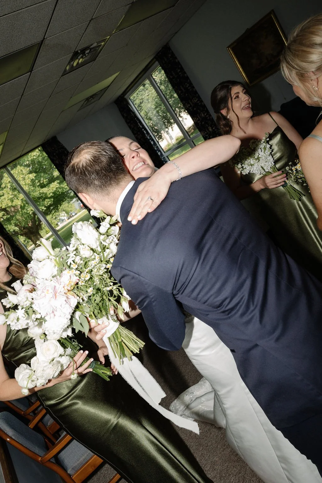 A wedding celebration where a bride and groom are hugging, surrounded by bridesmaids holding bouquets, inside a room with windows showing greenery outside.
