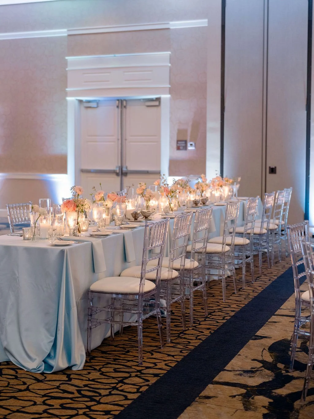 Elegant banquet table setup with white tablecloth, arranged with pink and white flowers, lit candles, and glassware, in a spacious room with beige walls and patterned carpet.