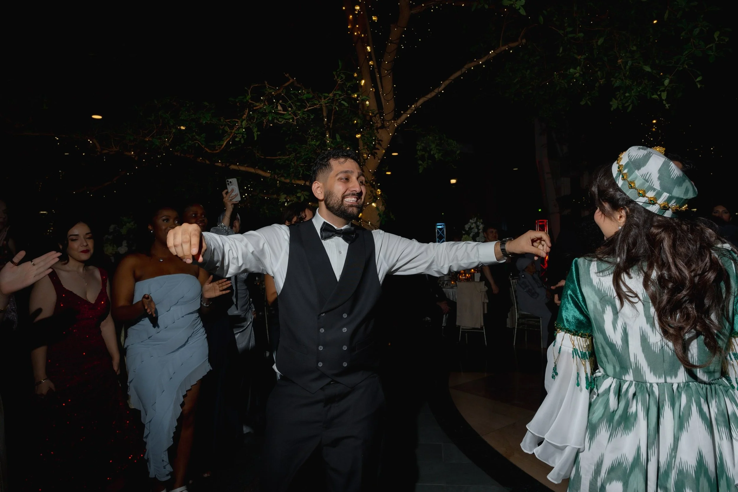 Man in tuxedo dancing at a celebration surrounded by women in formal dresses and a girl in a colorful dress wearing a decorative hat, under a decorated tree with lights at night.