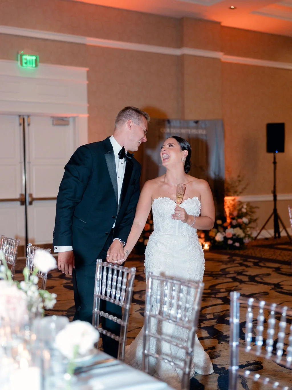 A newlywed couple sharing a happy moment at their wedding reception, holding hands and smiling joyfully, with the bride holding a glass of champagne.