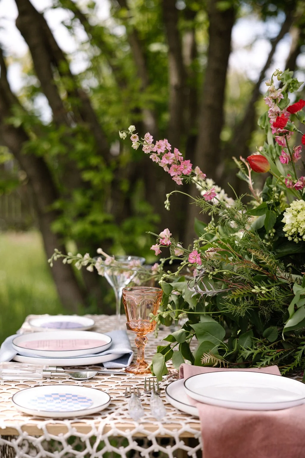 Outdoor table set with plates, glasses, and pink napkins, decorated with a lush floral arrangement of pink, white, and red flowers amidst green foliage, in front of a background of trees.