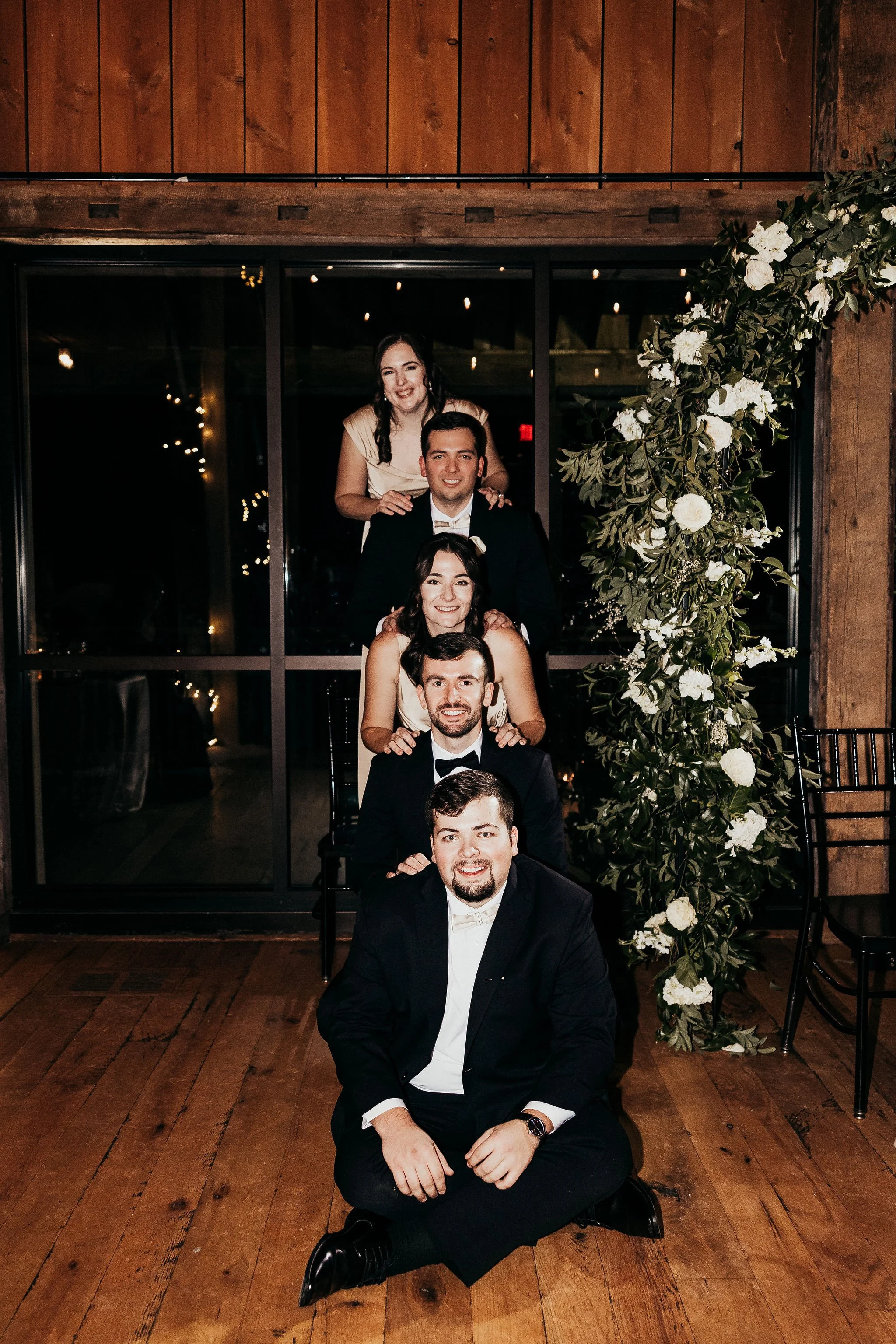A group of six people, dressed in formal attire, are stacked on top of each other in a line at an indoor event, with a decorative floral arrangement and a wooden wall in the background.